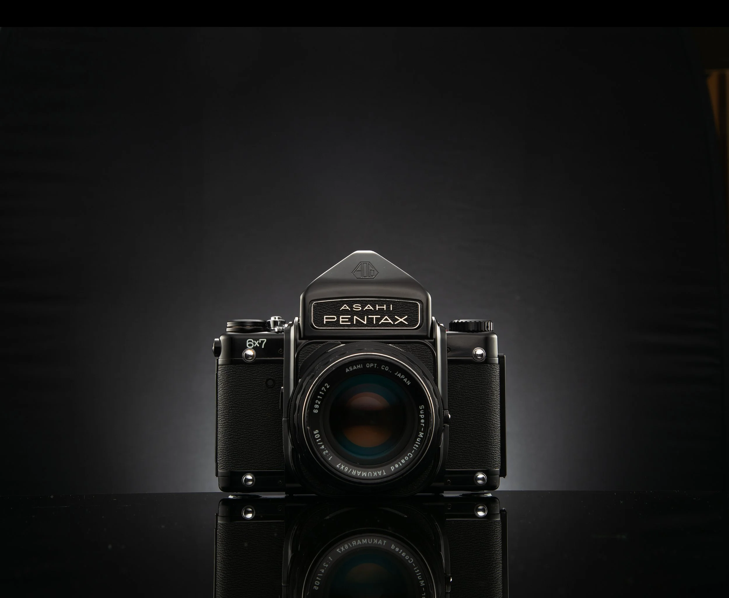 A vintage Asahi Pentax 35mm film camera with a 6x7 medium format lens, placed on a reflective black surface under focused lighting, with a dark background.