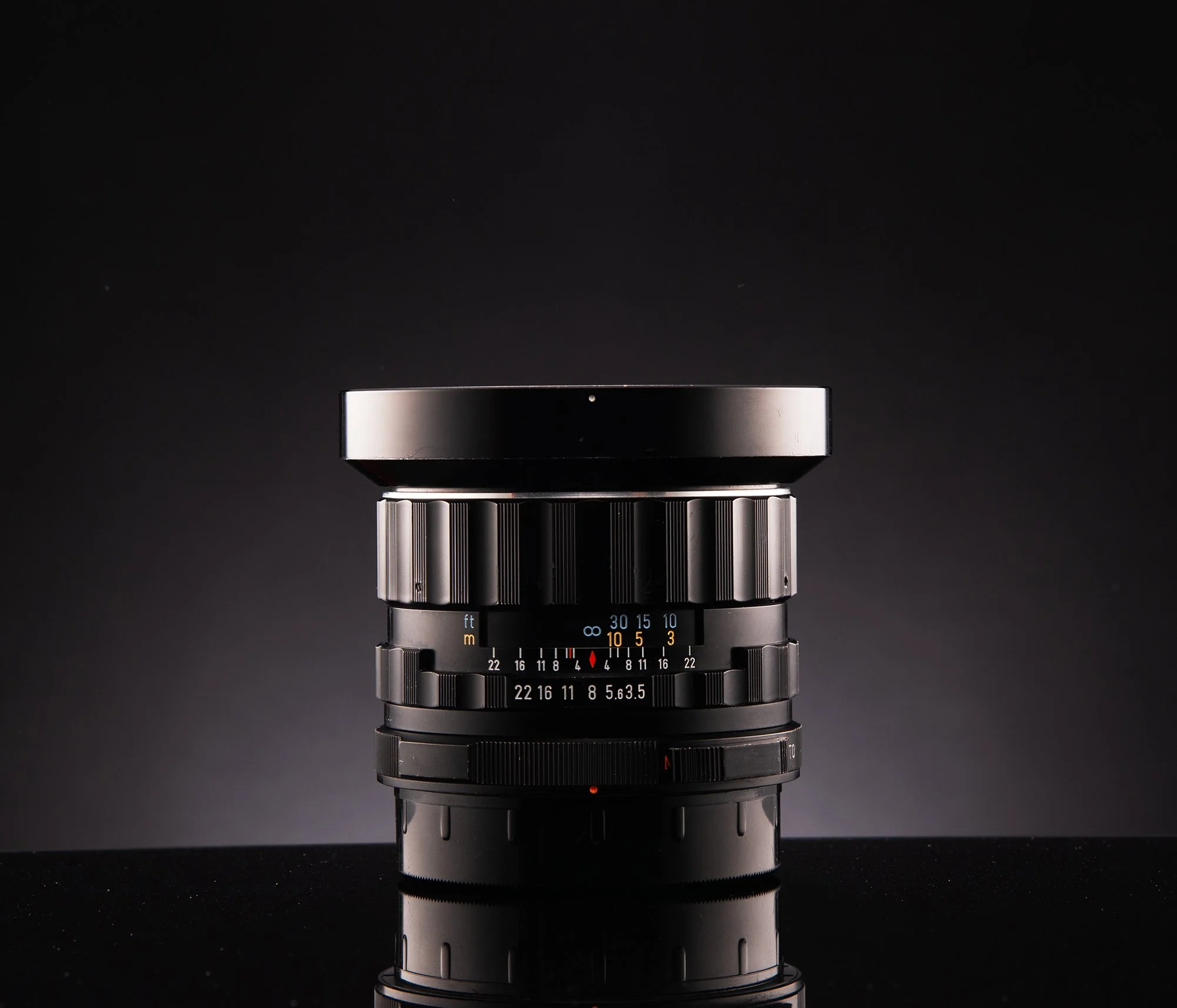 A black camera lens with various adjustment rings and lens markings, placed on a reflective black surface against a dark background.