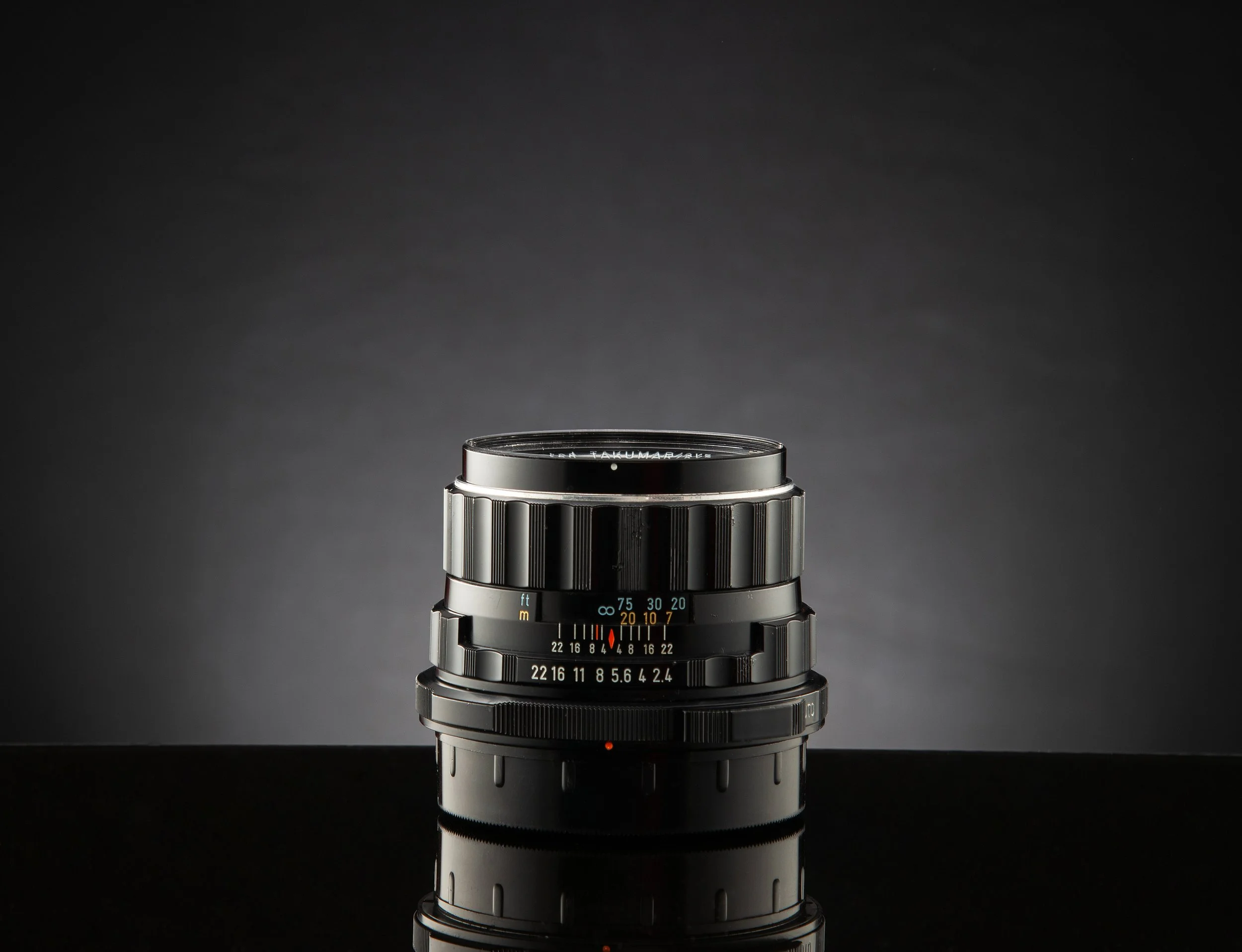 Close-up photo of a black camera lens against a dark gray background, reflecting on a glossy black surface.