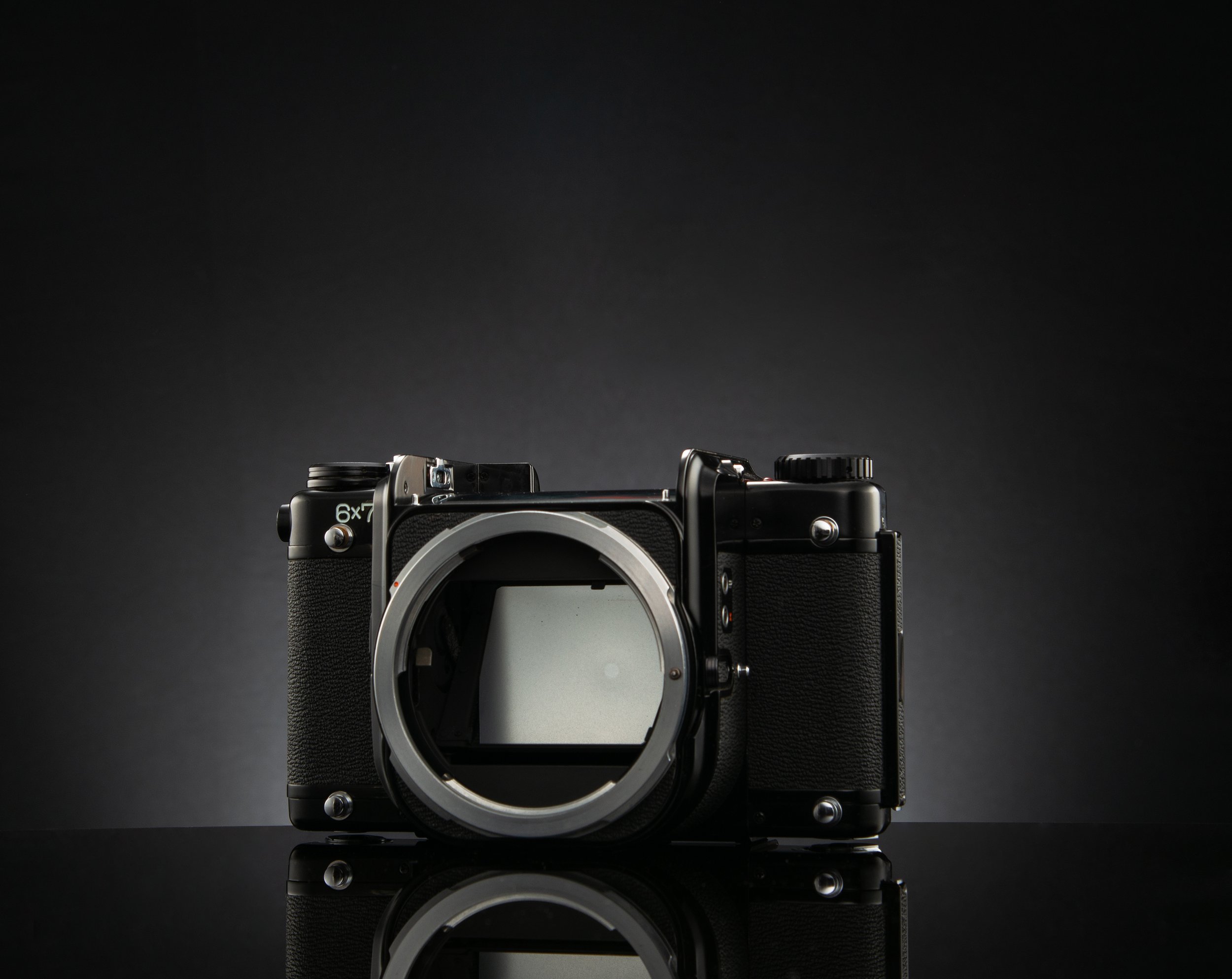 Asahi Pentax 6x7 body without a lens, placed on a reflective black surface against a dark gray background.