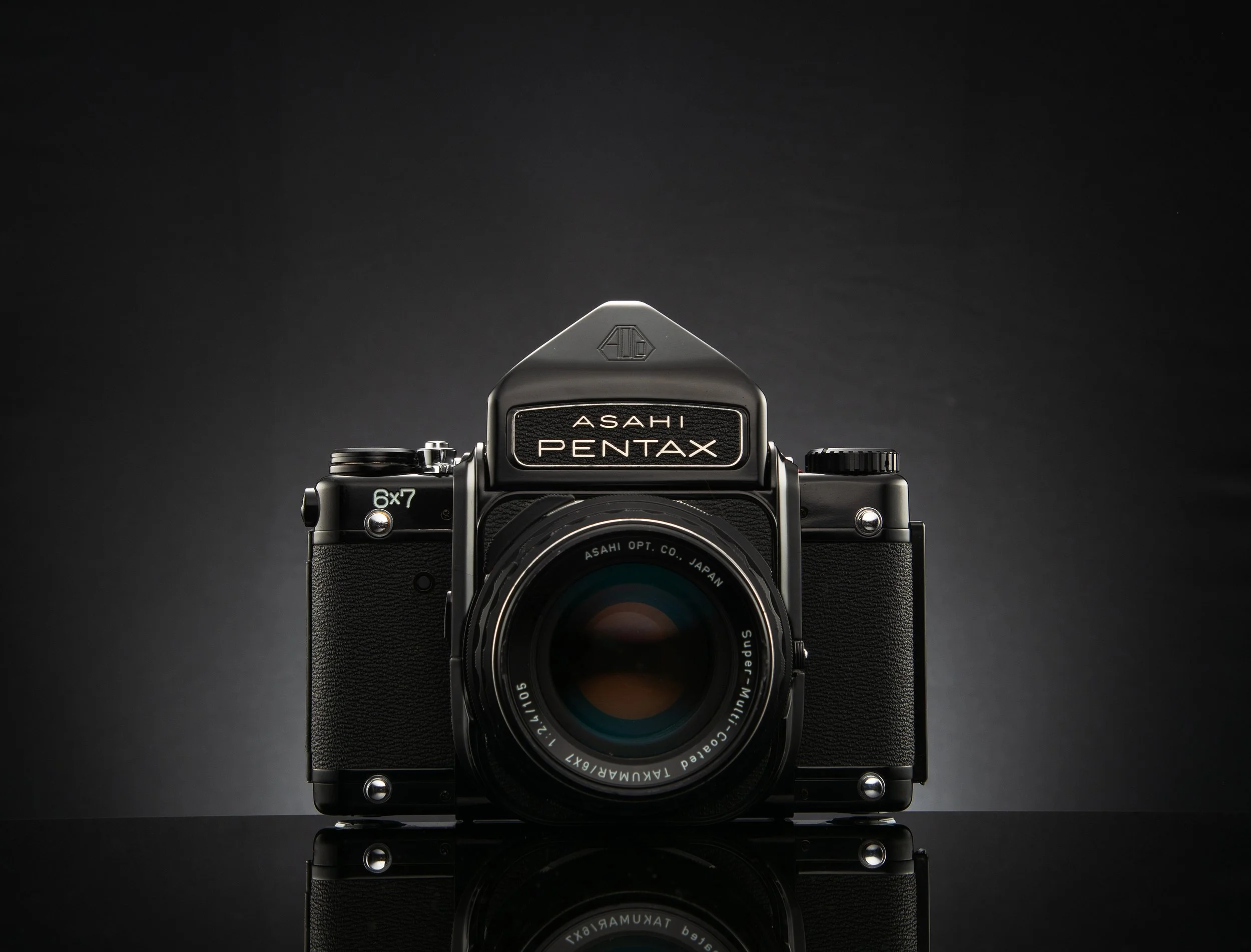 Asahi Pentax 6x7 camera with a lens, placed on a reflective surface, against a dark gradient background.
