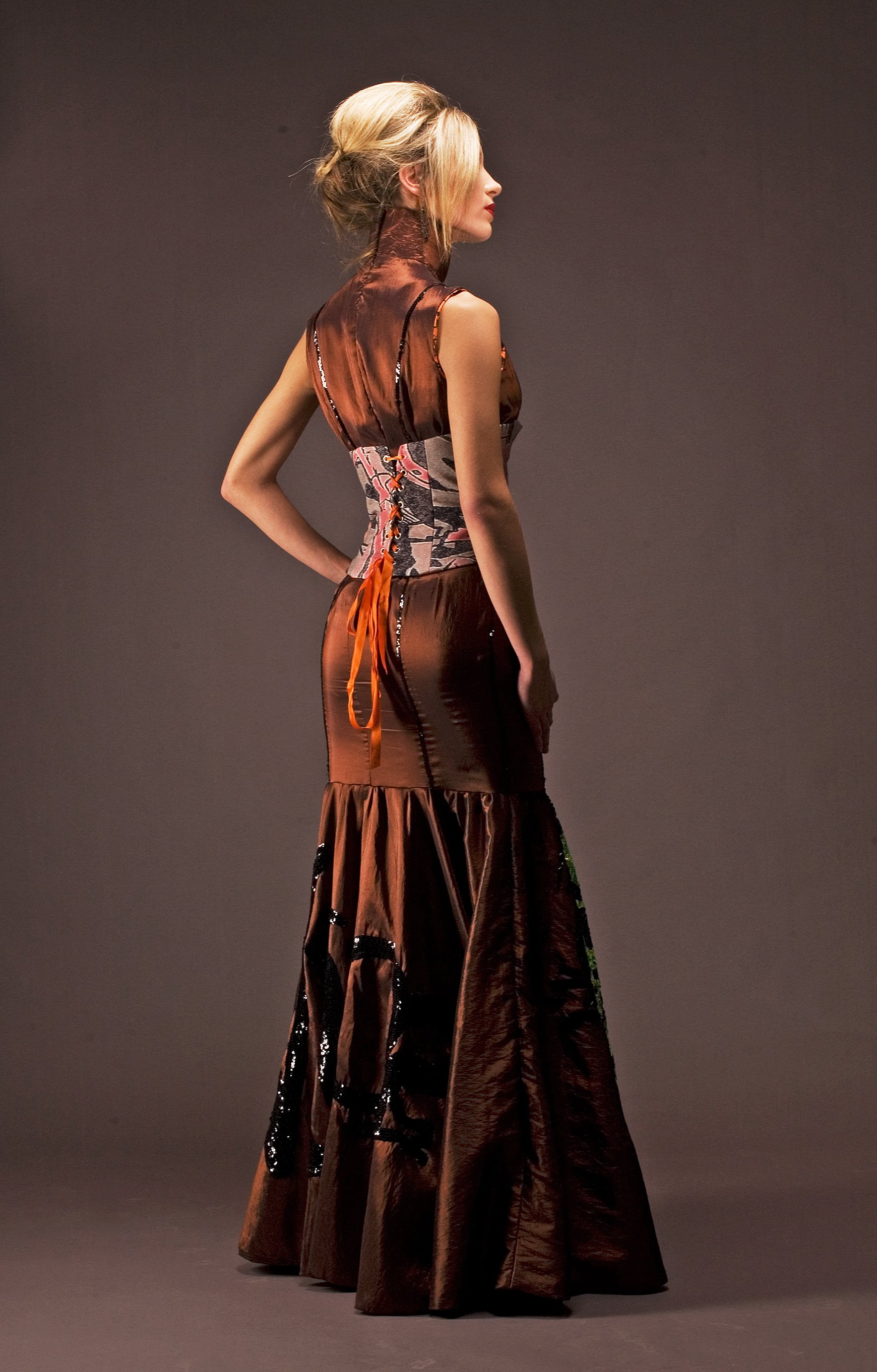 A woman in an elegant, brown, satin gown with ruffles, standing against a plain background, looking to the side.