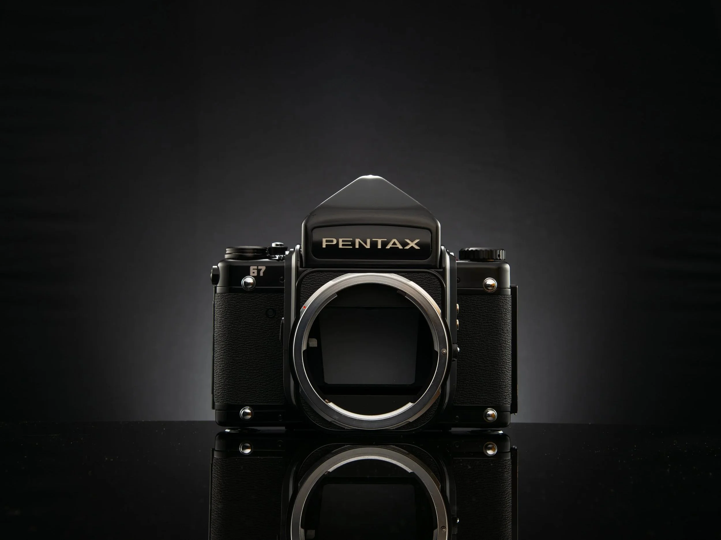 A black Pentax film camera with no lens mounted, set against a dark background with a reflective surface, illustrating a professional studio shot.