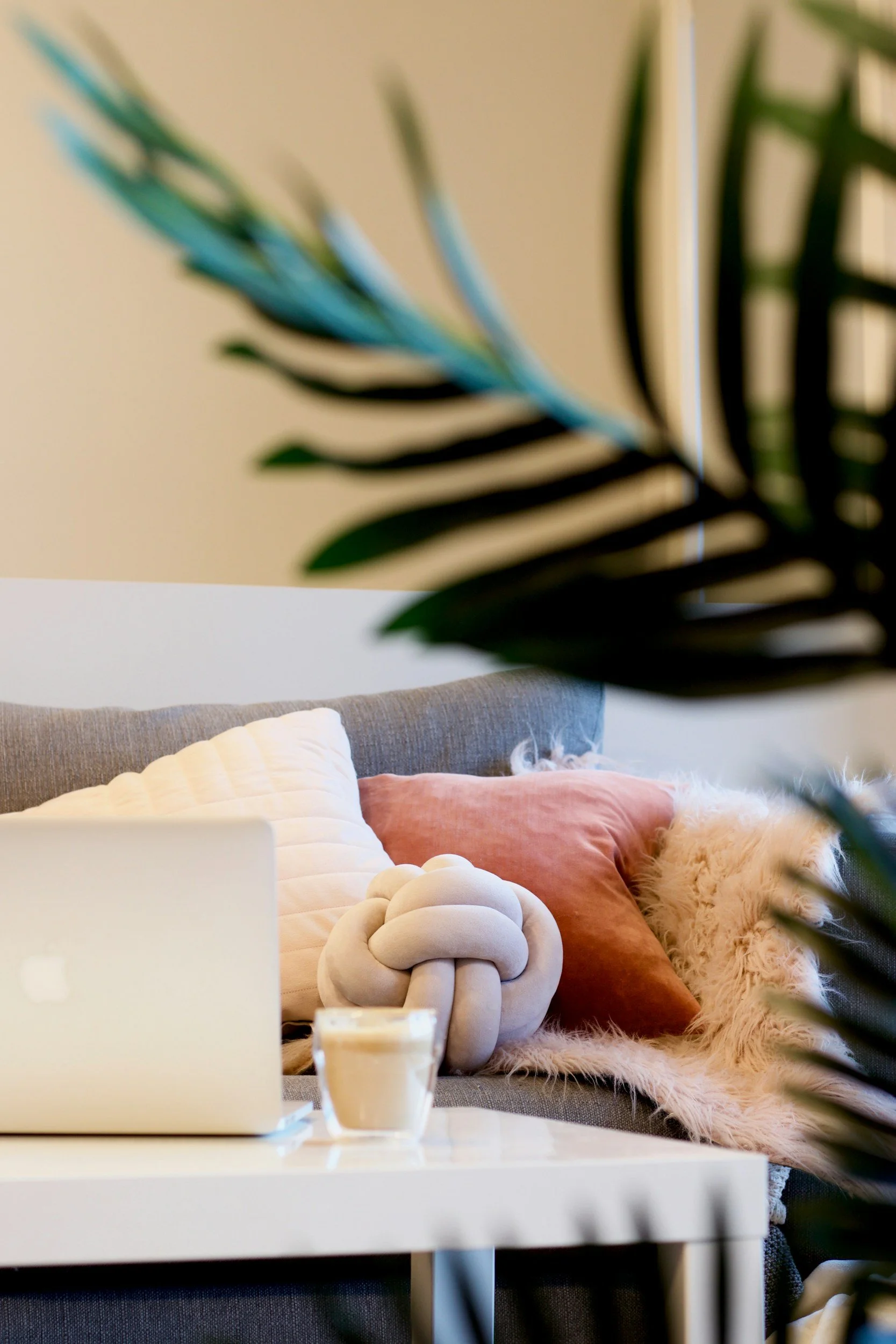 Cozy living room with beige walls, a gray couch decorated with white, blush pink, and fluffy beige pillows, and a knot-shaped beige cushion. On a white coffee table, there is a MacBook and a glass of latte. A green leafy plant partly blocks the view in the foreground.