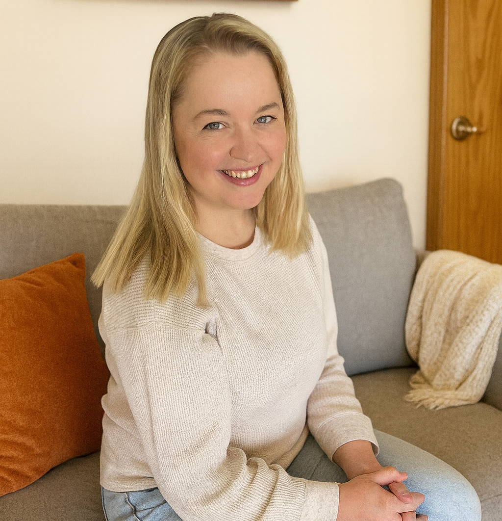 A woman with blonde hair sitting on a gray couch, smiling, wearing a cream sweater and light jeans in a cozy living room.