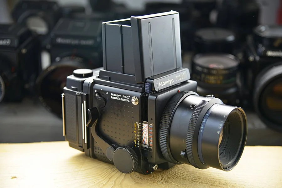 A Mamiya RZ67 PROFESSIONAL camera with a large lens and film magazine on a wooden surface, backdropped by various camera lenses.