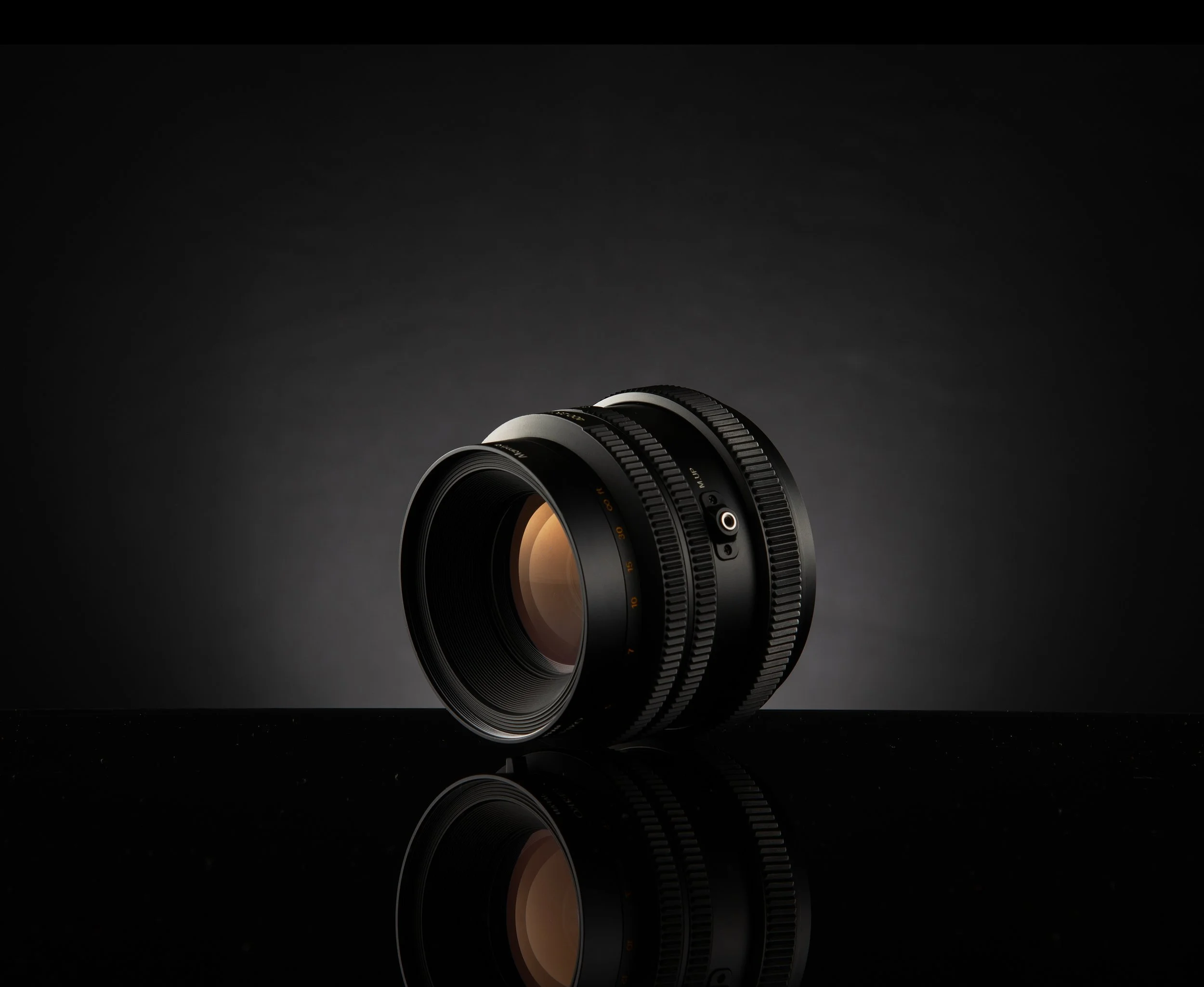 A camera lens resting on a reflective surface against a dark gradient background.