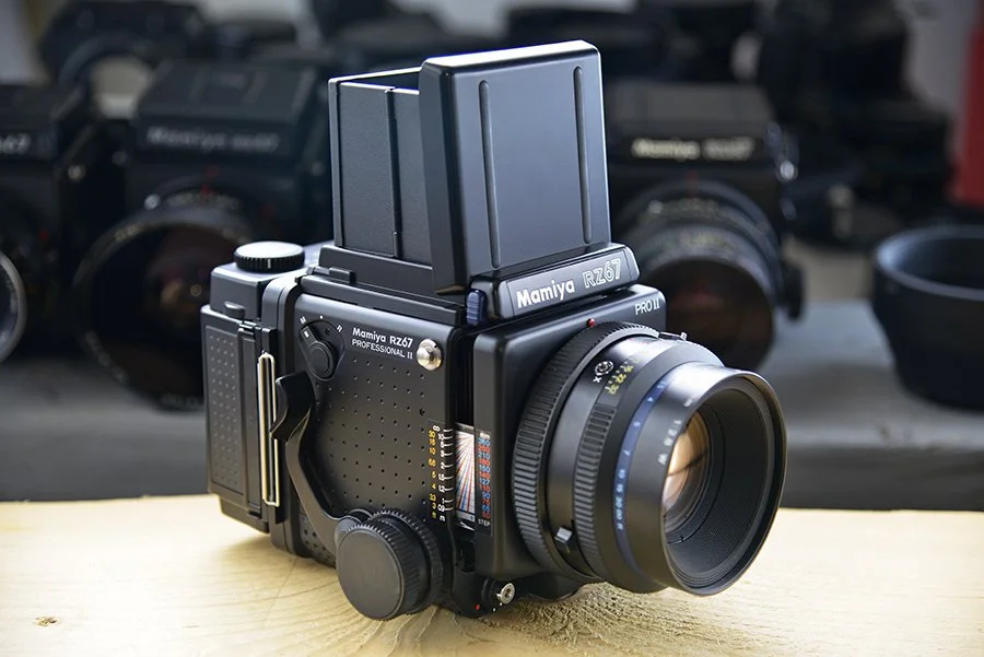 Mamiya RZ67 professional medium format camera with waist-level viewfinder on a wooden surface, with other camera equipment in the background.