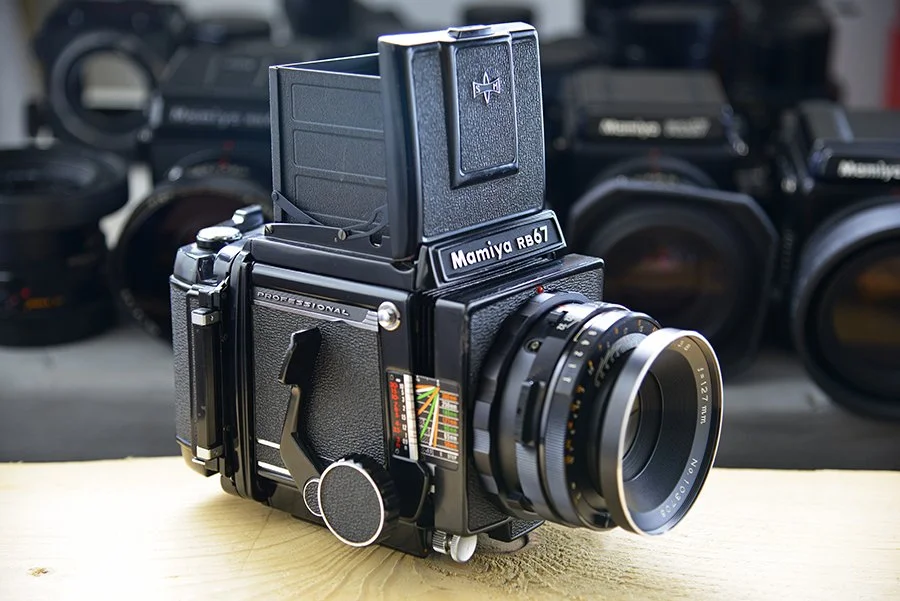 A vintage Mamiya RB67 medium format film camera with a lens attached, placed on a wooden surface with camera equipment blurred in the background.
