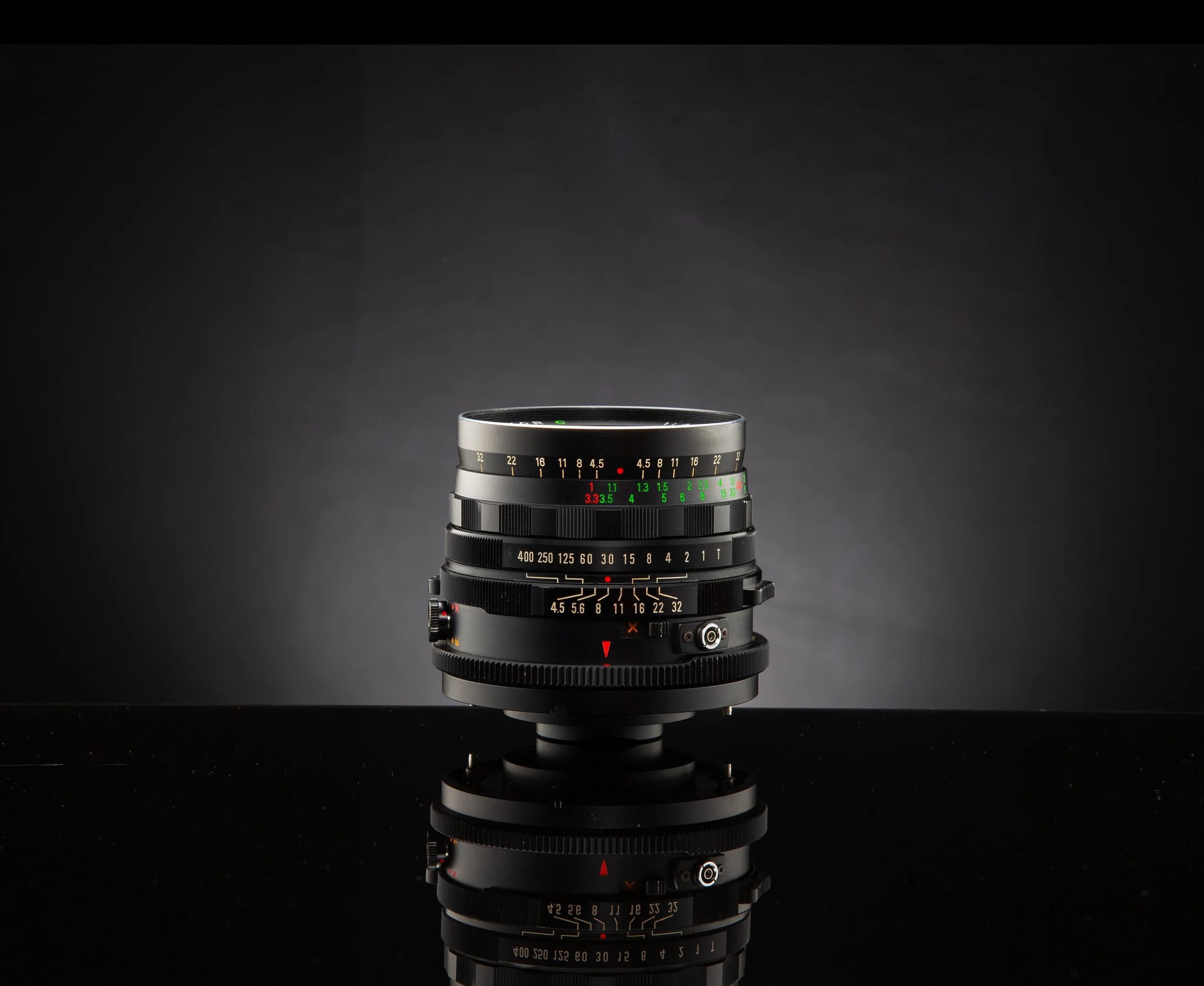 A close-up of a vintage camera lens with various focus and aperture markings, placed on a reflective black surface against a dark background.