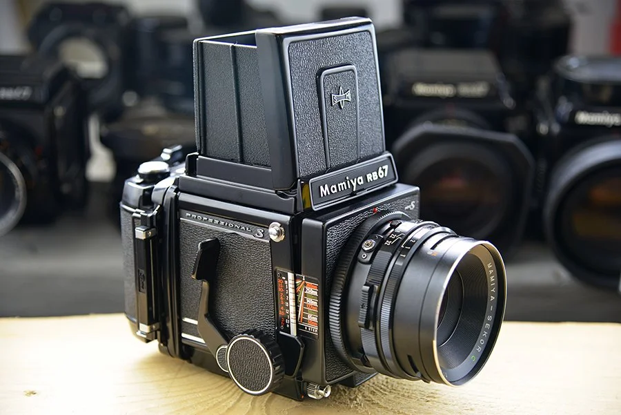 A vintage Mamiya RB67 medium format camera with a detachable viewfinder, placed on a wooden surface, with other cameras blurred in the background.