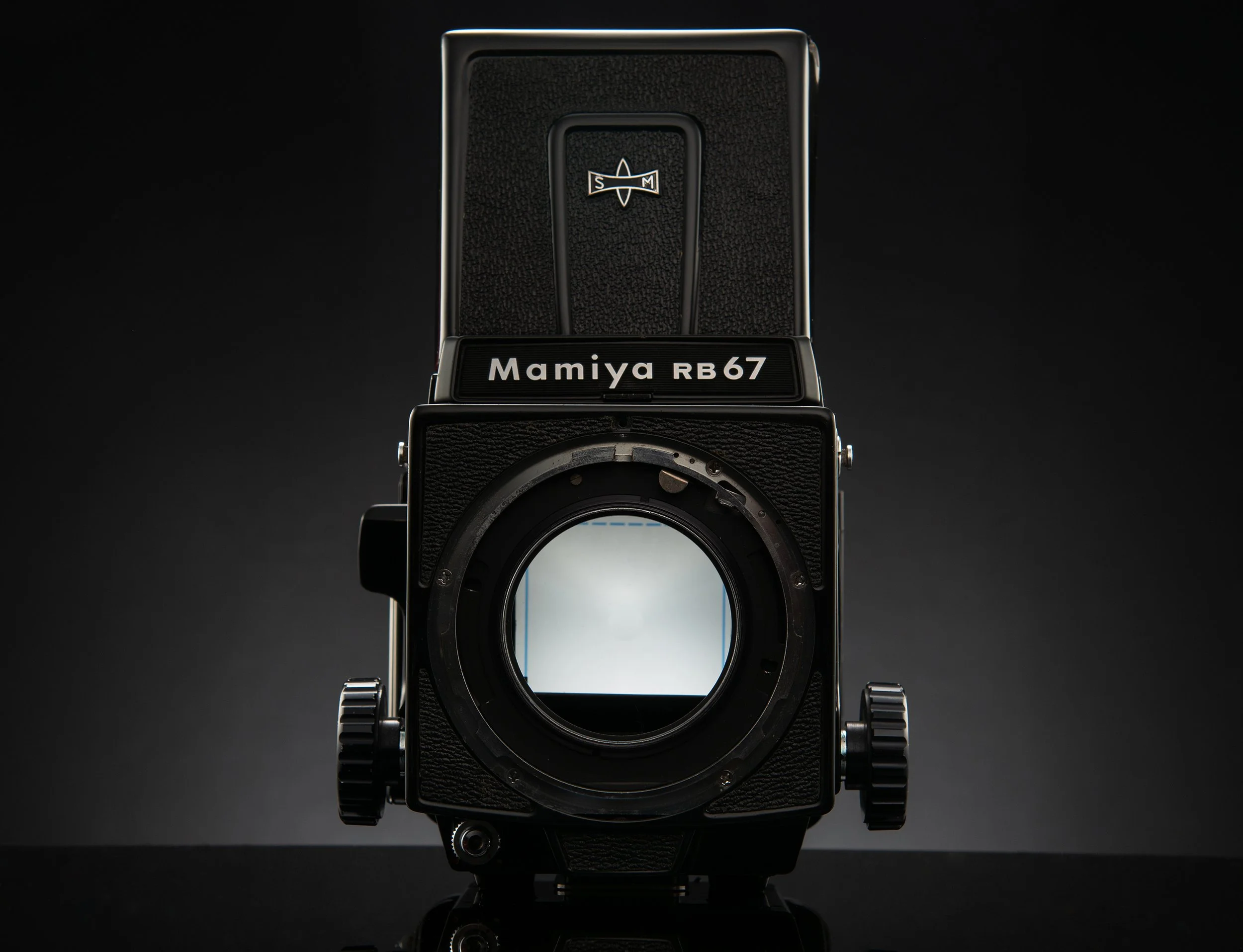 A Mamiya RB67 medium format camera with a black body, lens removed showing the mirror inside, against a dark background.