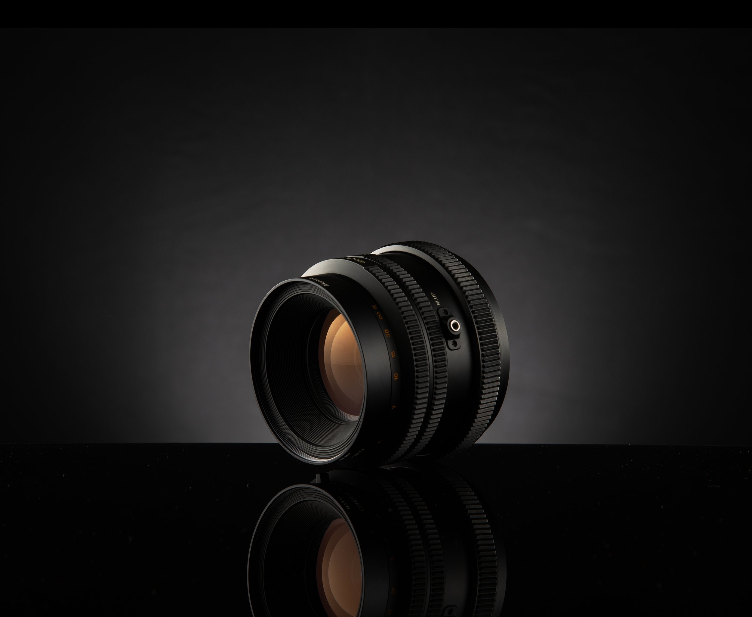A camera lens against a dark gradient background with a reflective surface below.