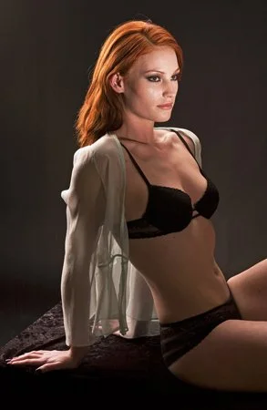 A woman with red hair wearing black lingerie and a sheer green shirt, sitting against a dark background.