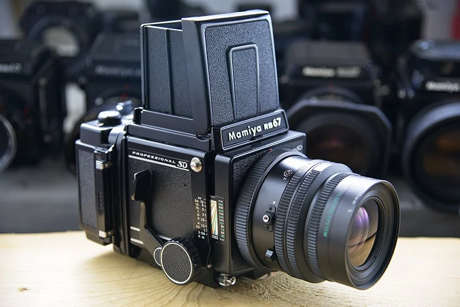 A professional medium format camera with a large lens, labeled 'Mamiya RB67,' placed on a wooden surface with multiple other cameras in the background.