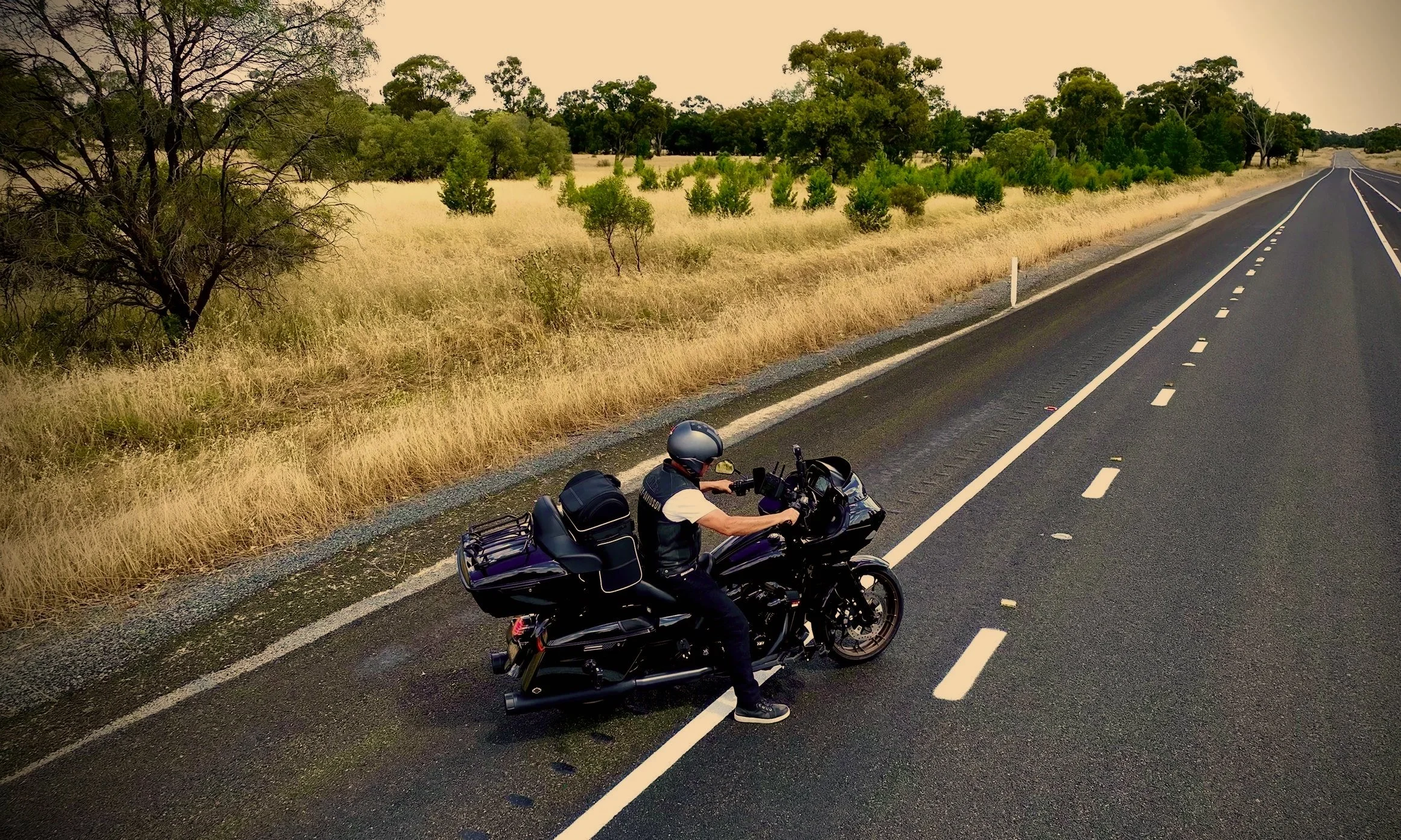 Motorcyclist stopped on the side of a rural highway with trees and grass in the background. The highway is empty, vast and very long.