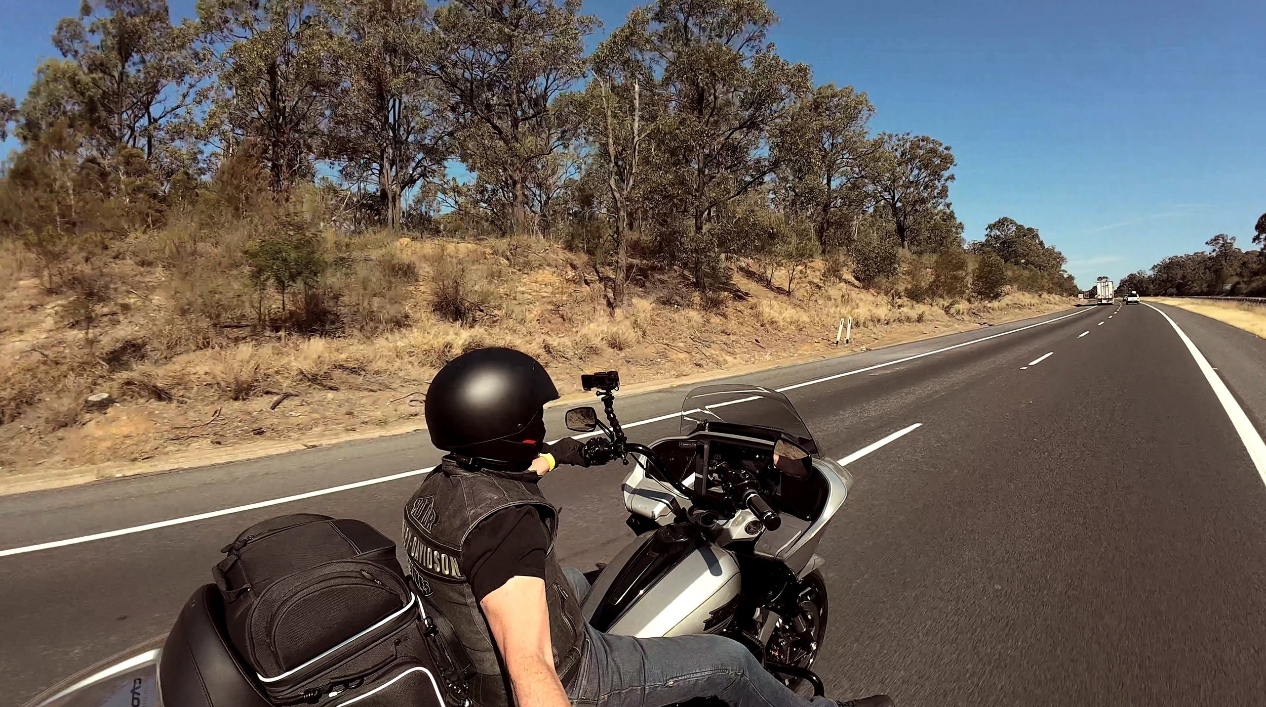 A motorcyclist wearing a black helmet and leather vest riding a motorcycle on a highway with dry grass and trees on the side under a clear blue sky.