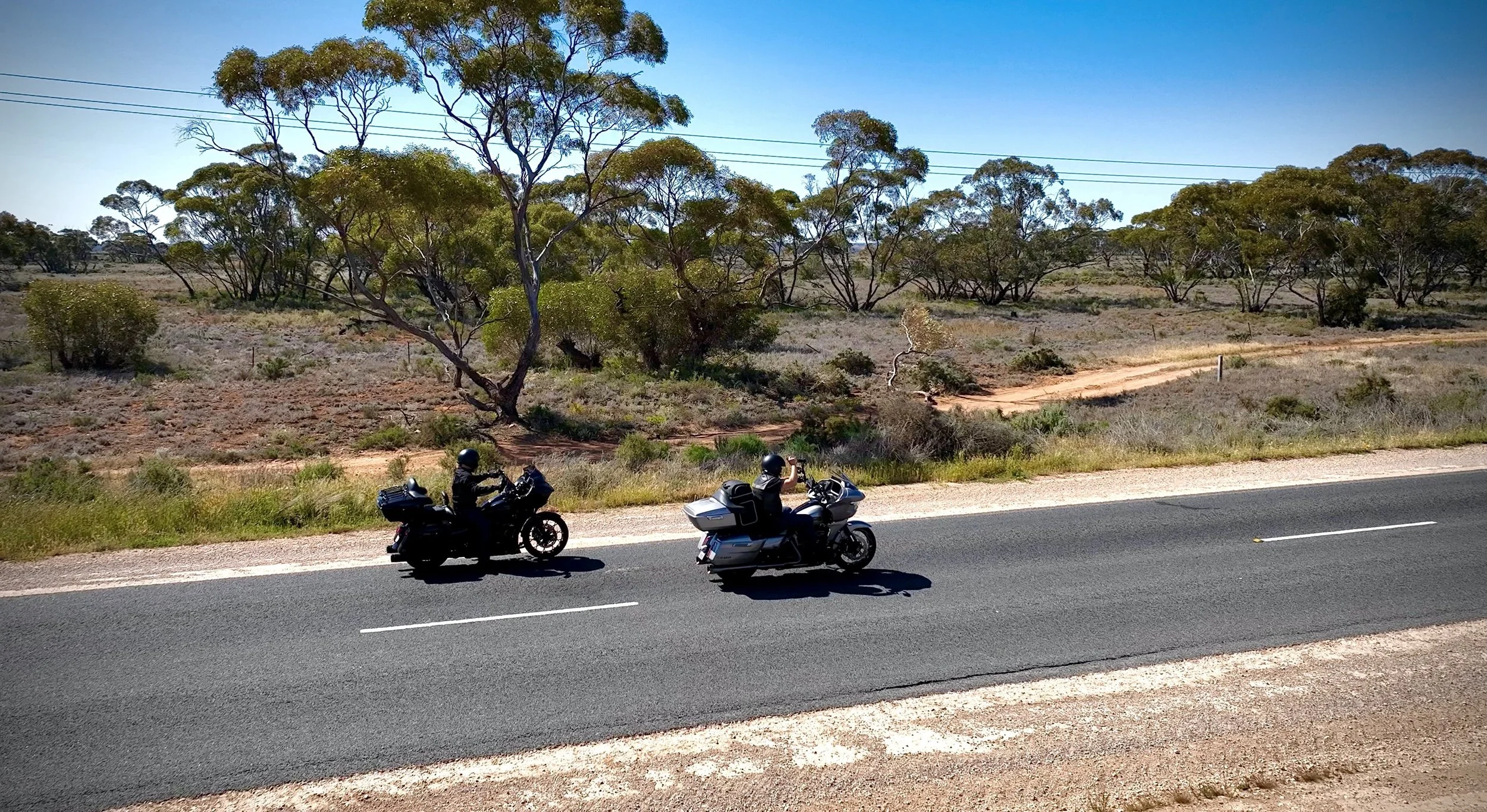 Two motorcyclists wearing helmets riding on a paved road through a dry, open landscape with trees and bushes.