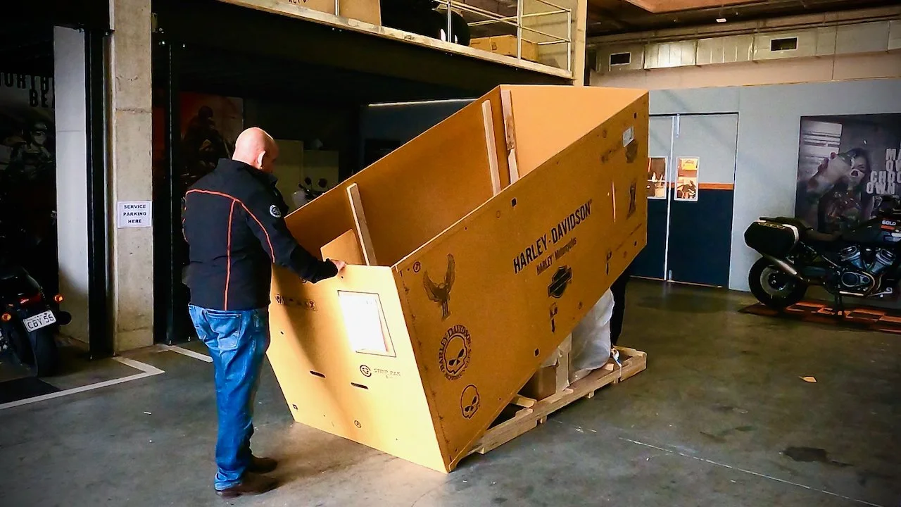 A man inspecting a large yellow wooden crate with Harley-Davidson and skull logos in an indoor space, with bikes nearby and a sign that says "Service Parking Here." He is unboxing a new motorcycle.