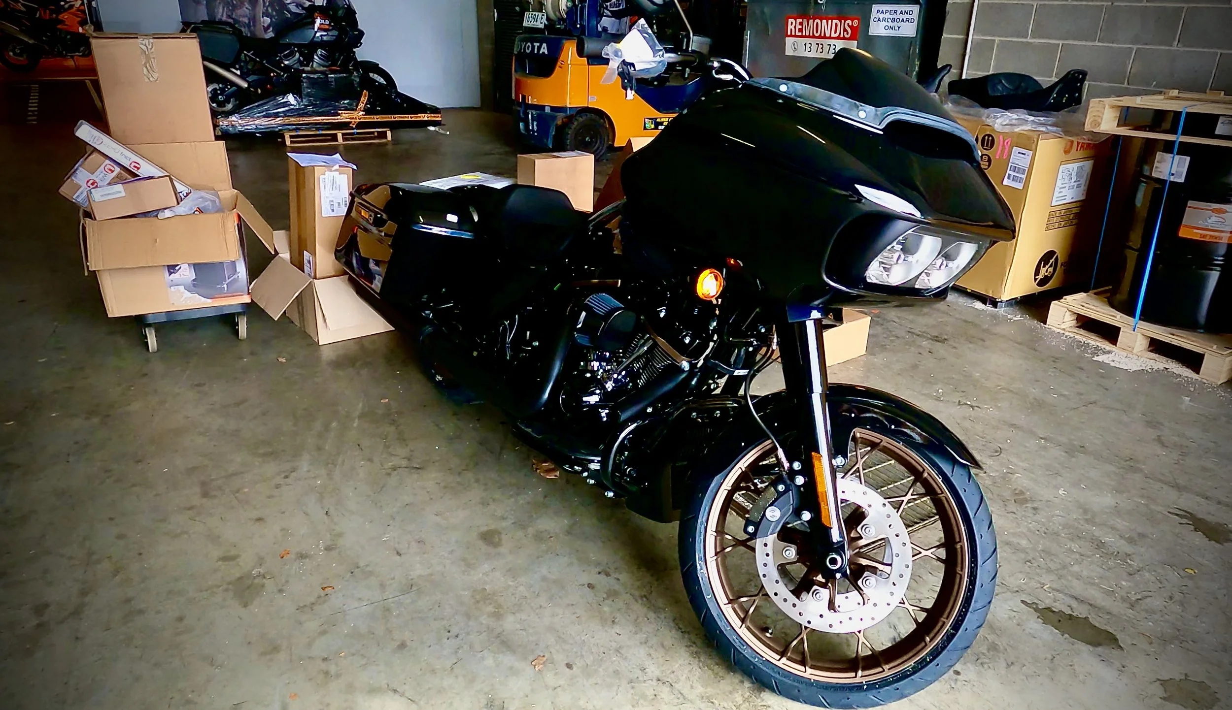 A black touring motorcycle in a warehouse or storage area, surrounded by cardboard boxes, pallets, and many motorcycle parts.