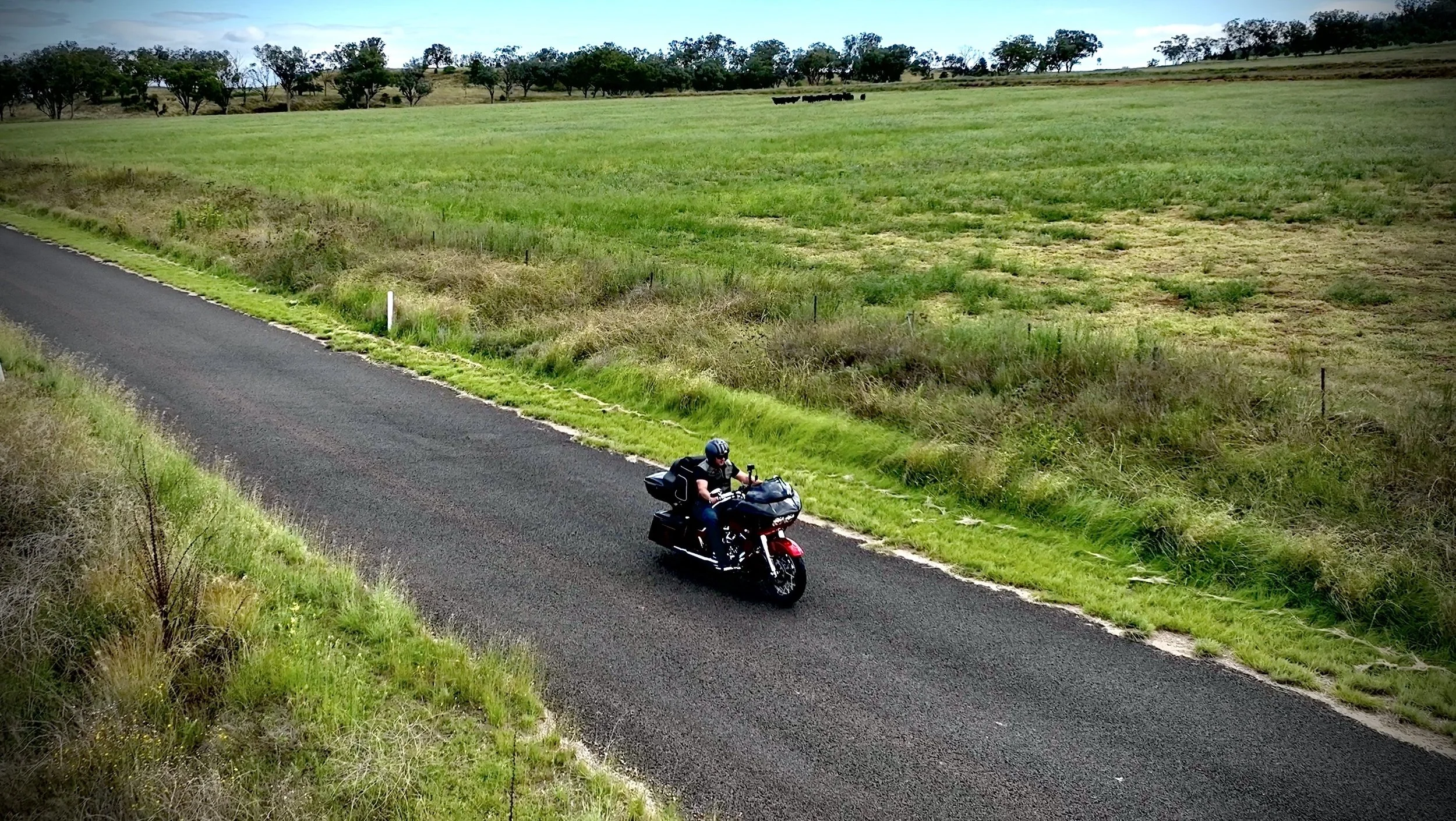 A person riding a black motorcycle on a rural road with green fields and trees in the background.