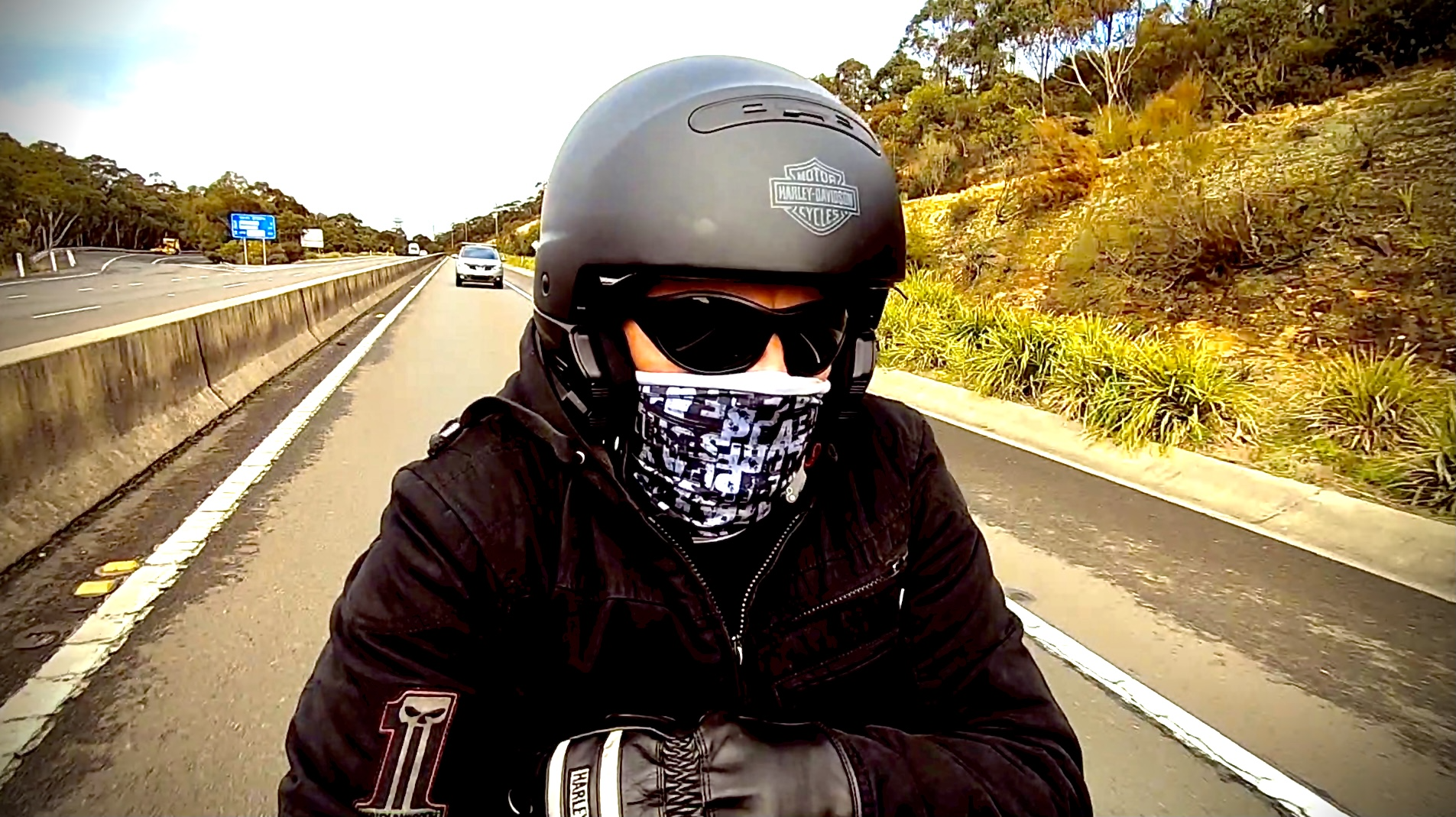 A motorcyclist wearing a black helmet, sunglasses, a patterned face mask, a black jacket, and gloves riding on a road with cars passing by, with trees and hillside on the side.