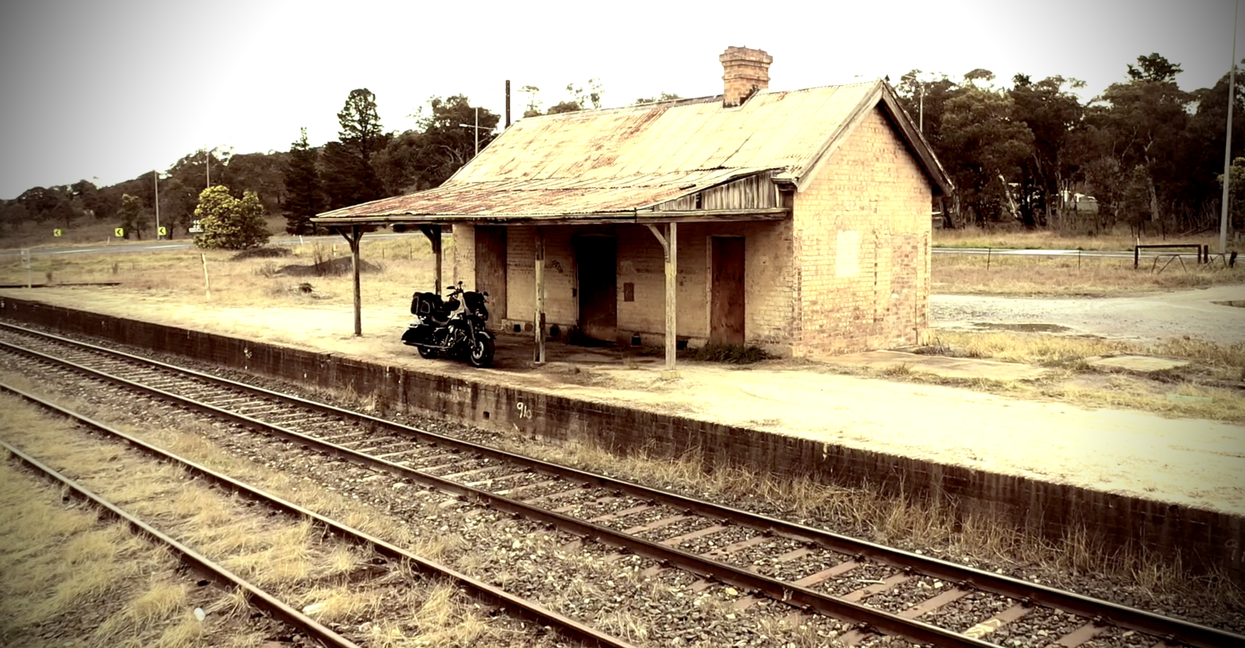 An old brick train station building with a rusty metal roof and wooden pillars, situated alongside train tracks with a motorcycle parked on the station platform, in a rural setting with trees in the background.