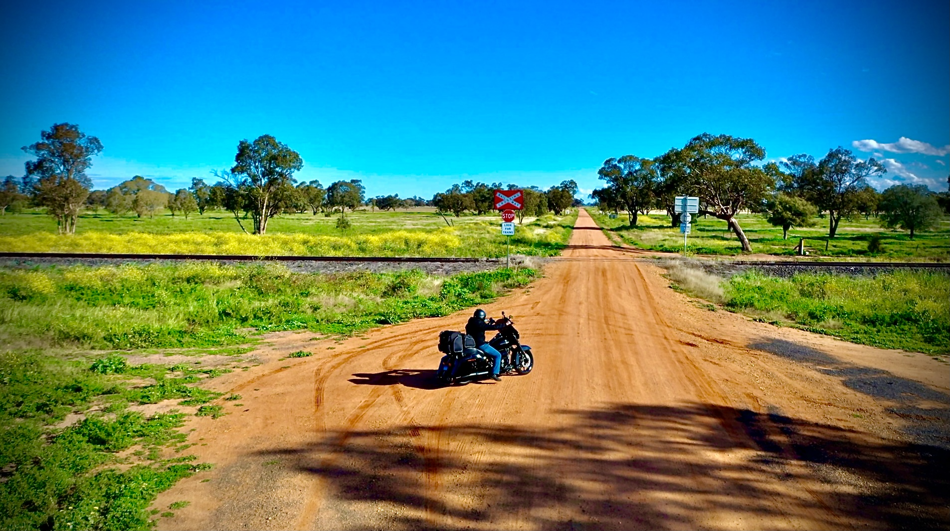 A person riding a black motorcycle on a dirt road with green fields and trees in the background, crossing railroad tracks on an orange dusty road on a sunny day.