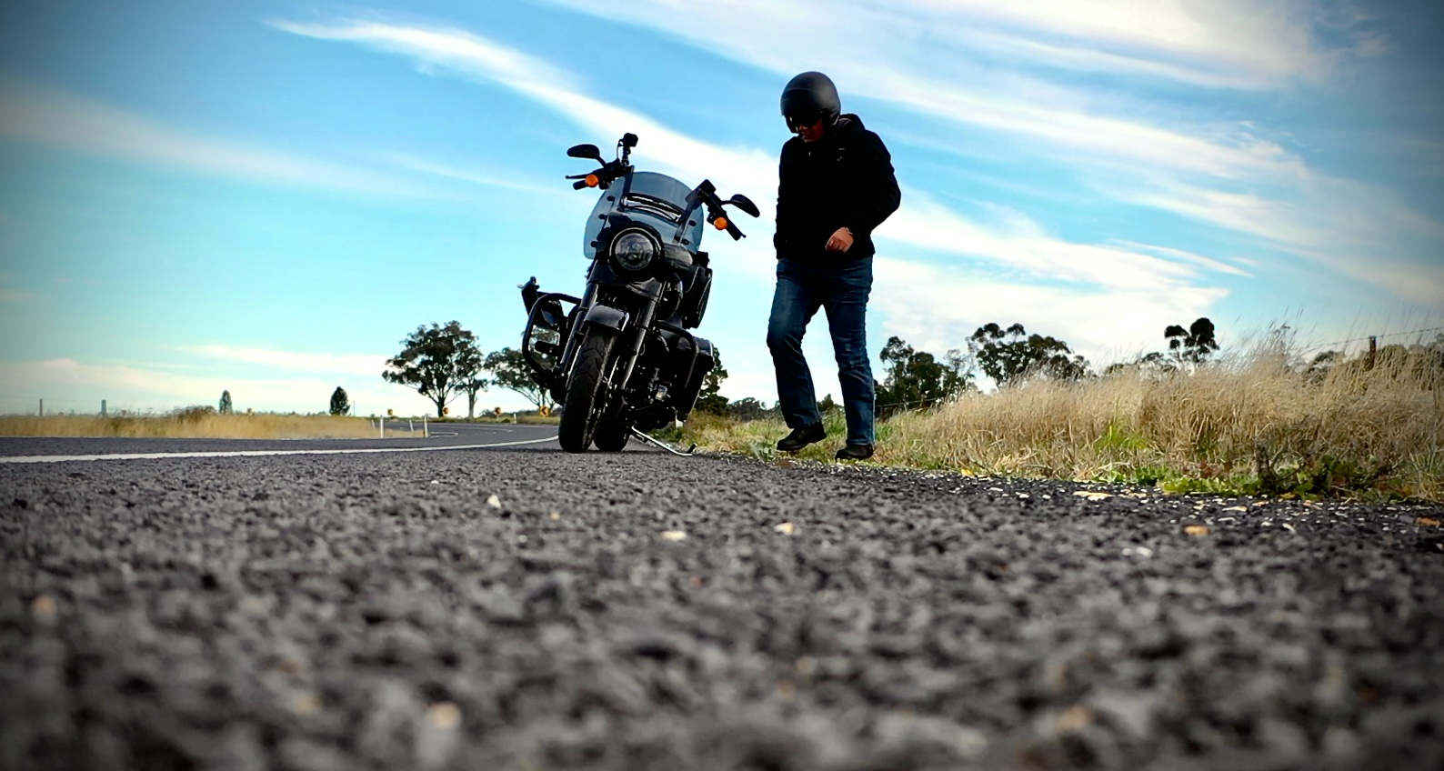Person in black jacket and jeans getting off a black motorcycle parked on the side of a rural road with grass and trees in the background under a partly cloudy blue sky.
