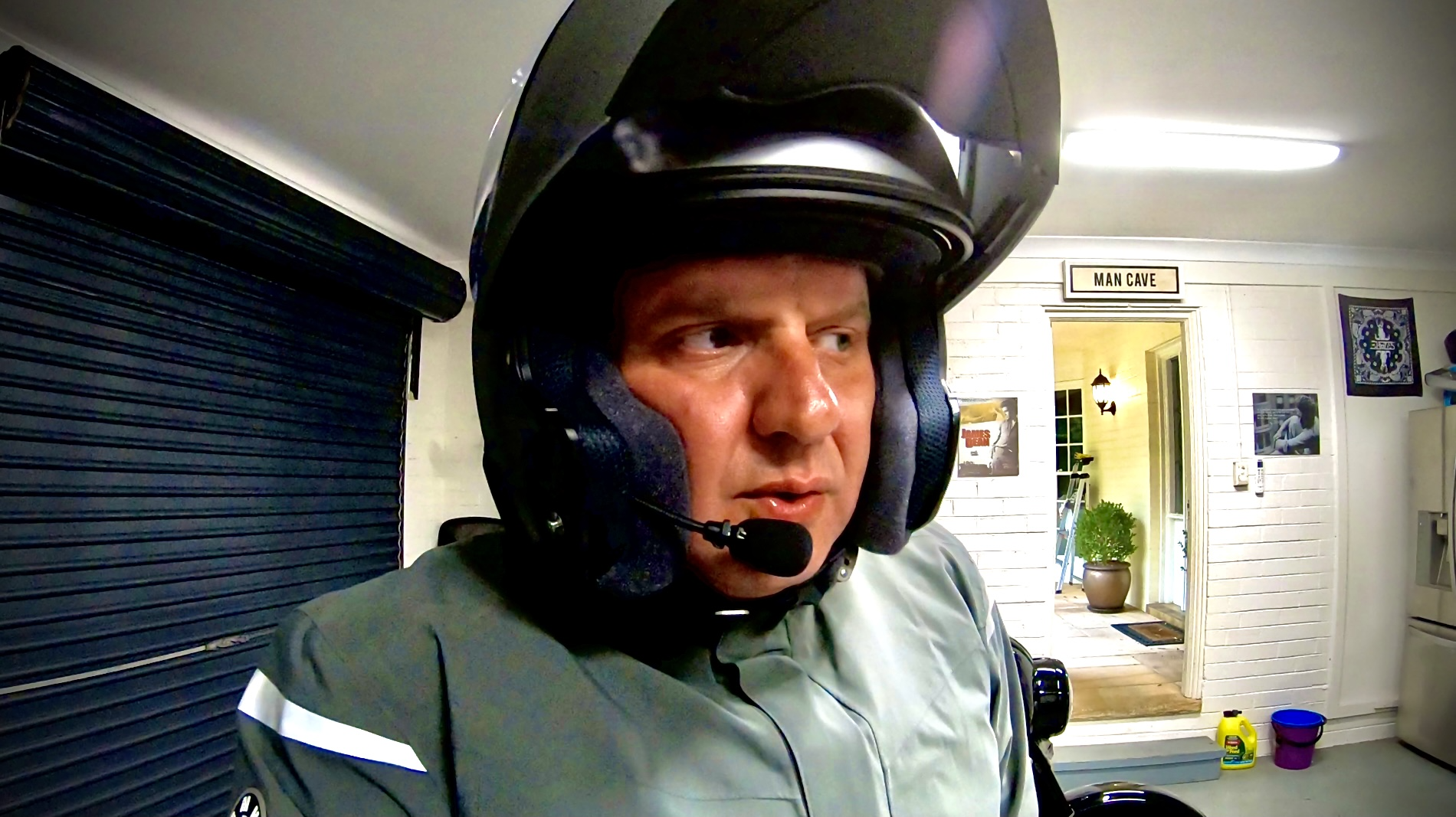 A man wearing a motorcycle helmet with a communications microphone attached, in a garage or indoor space. There is a sign that says, 'Man Cave'.