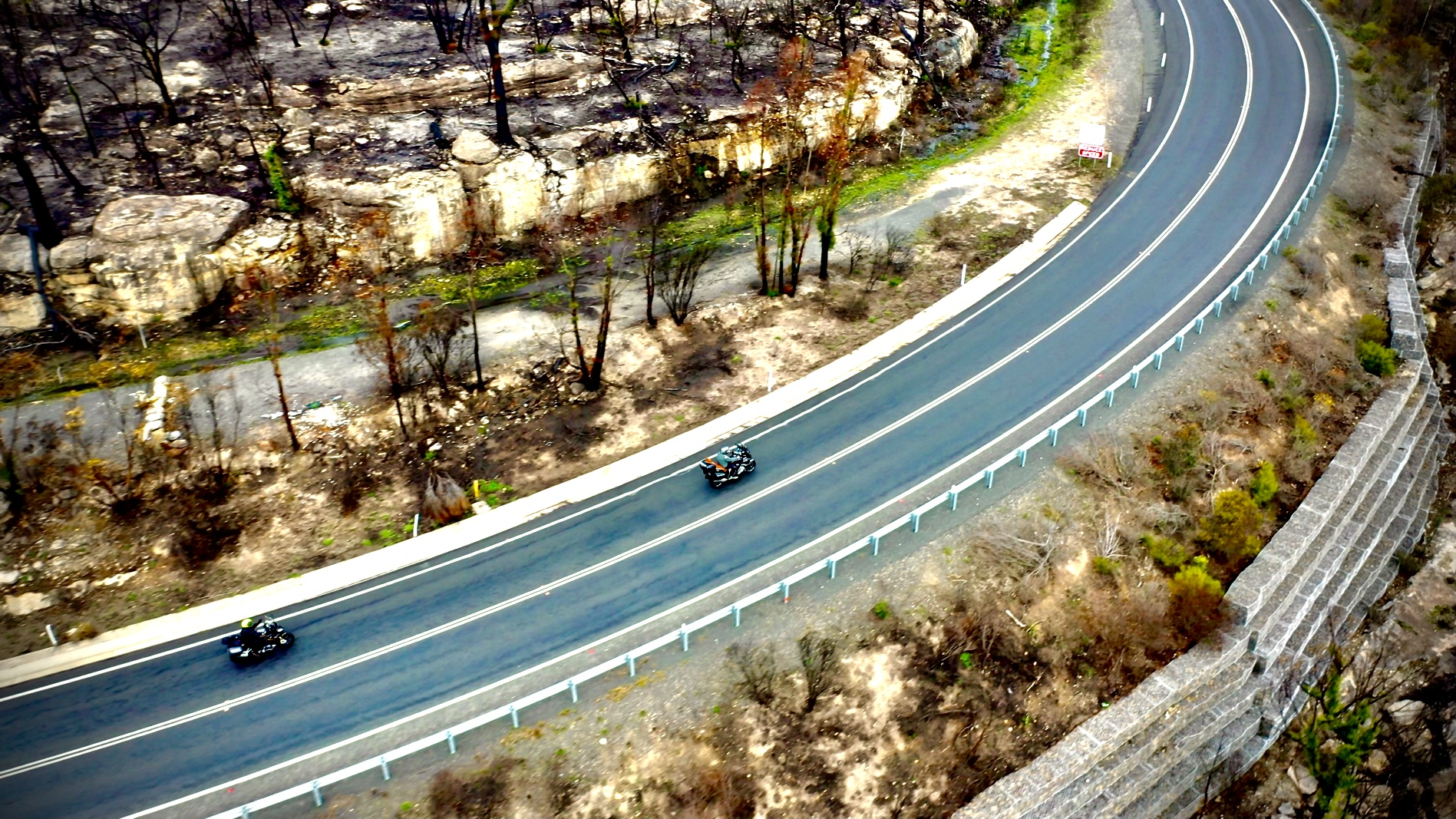 Two motorcycles traveling on a winding mountain road with a rocky hillside and sparse trees on one side and a stone retaining wall on the other.