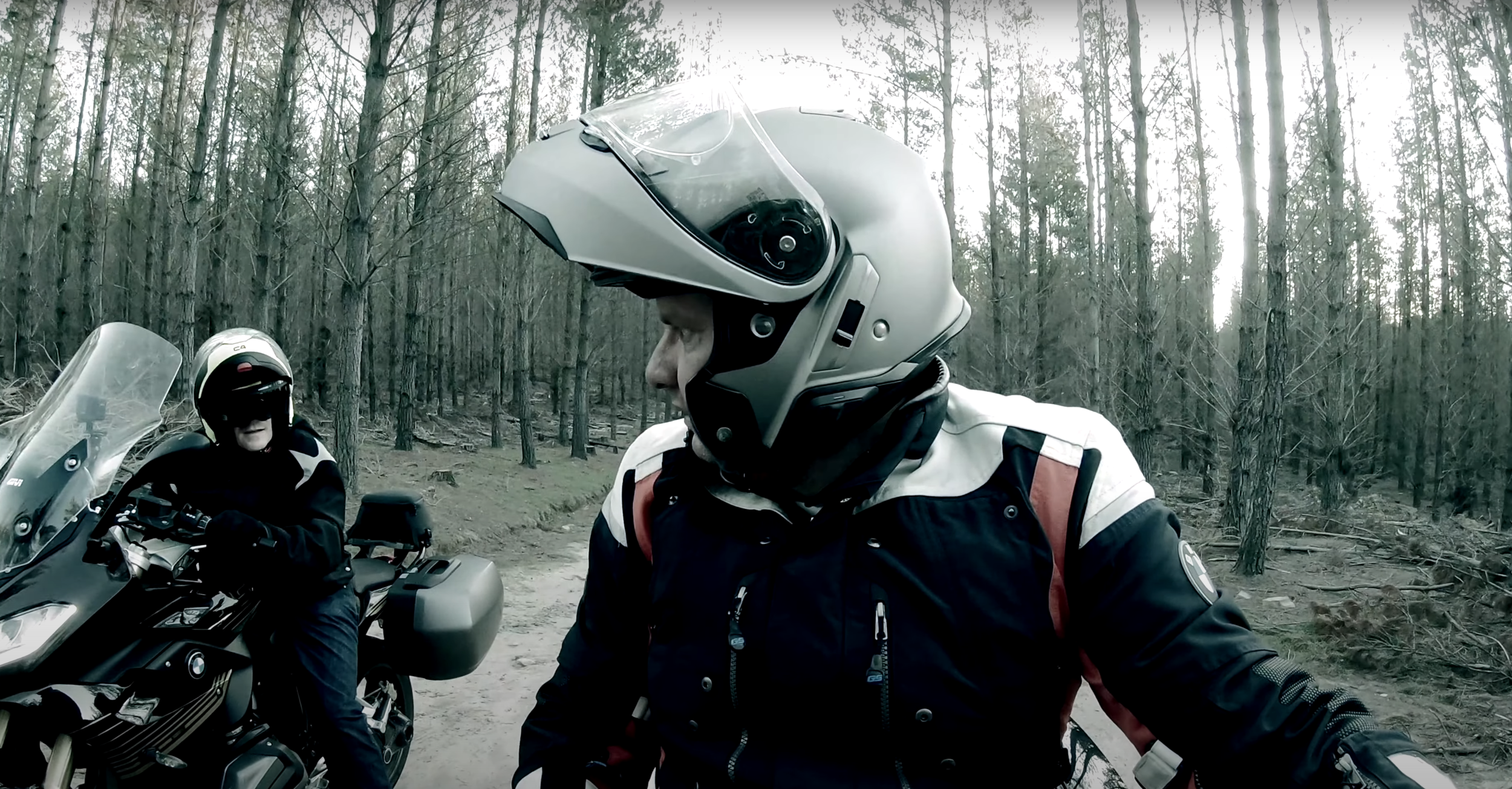 Two motorcyclists wearing helmets and riding gear in a dark forested area with tall trees and a dirt trail.