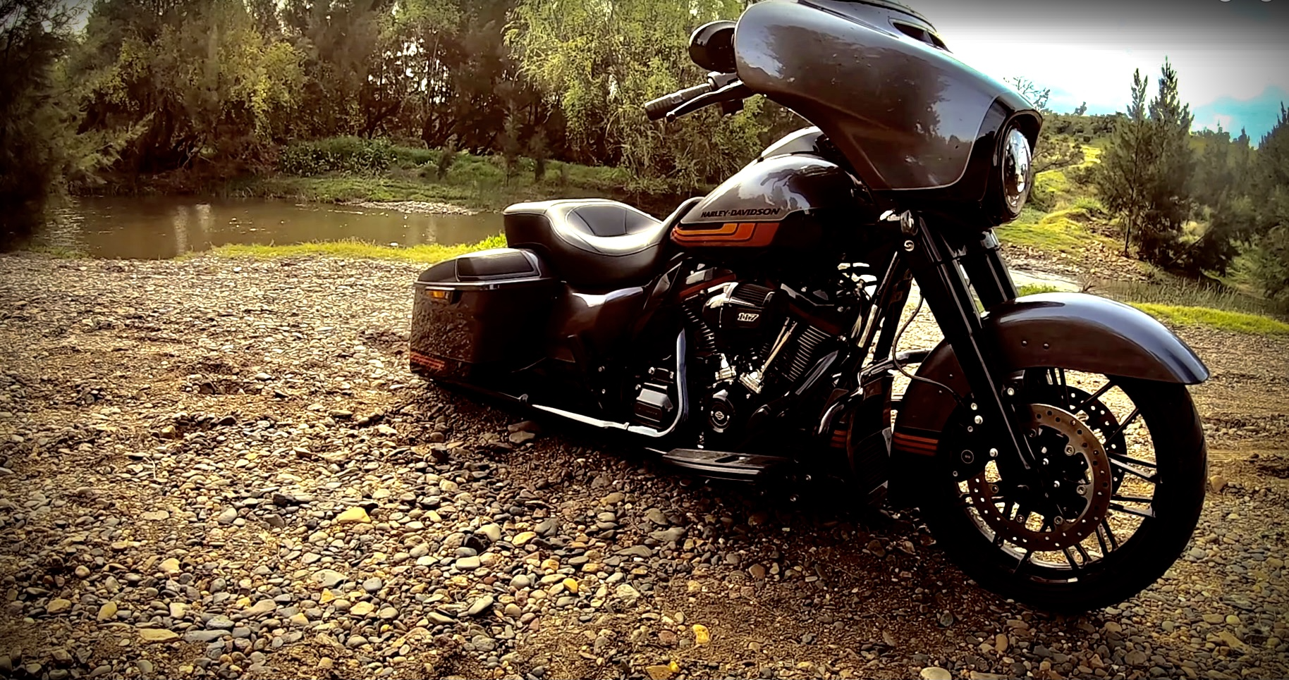 A Harley Davidson touring motorcycle bogged on a rocky riverbank with trees and a small river in the background.