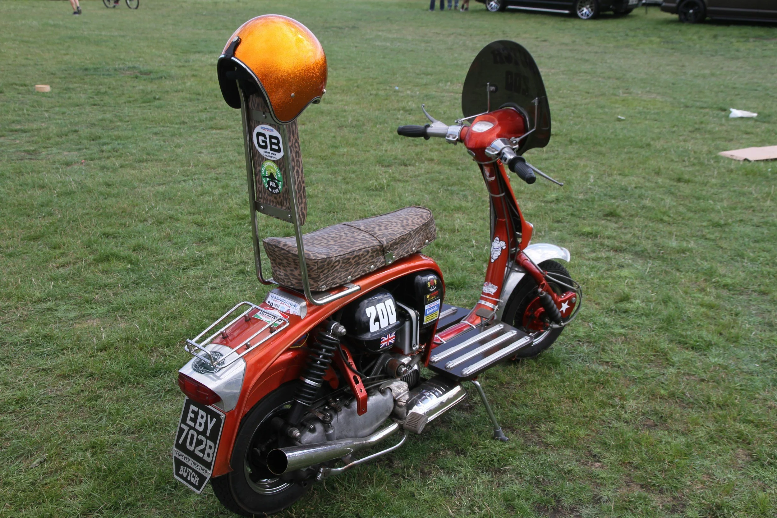 A small vintage red scooter with a leopard print seat, a short windshield, and a rear luggage rack, parked on grass with some cars in the background.