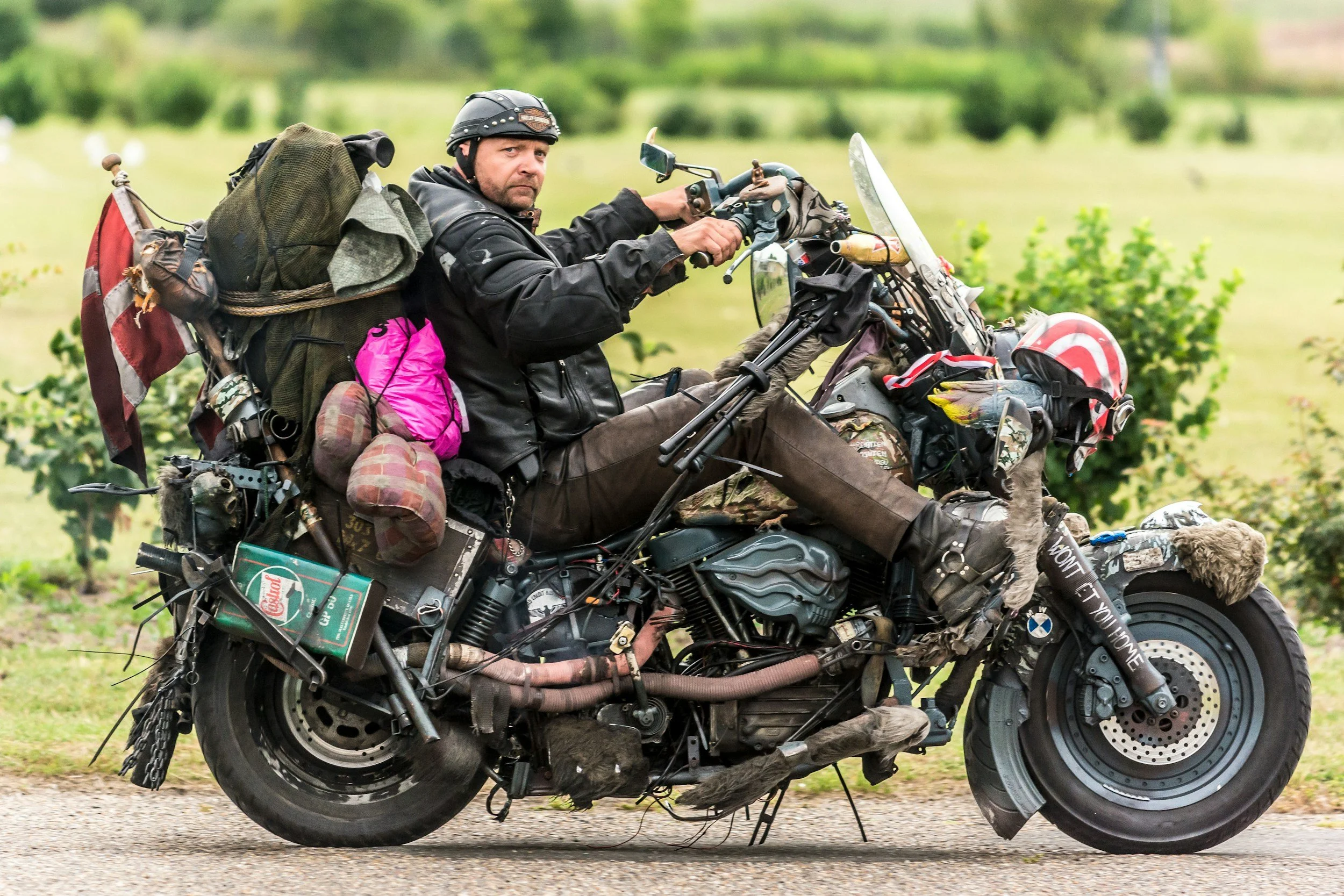 A man in motorcycle gear riding a heavily customized motorcycle, carrying large bags and equipment, on a rural road with green grass and trees in the background.