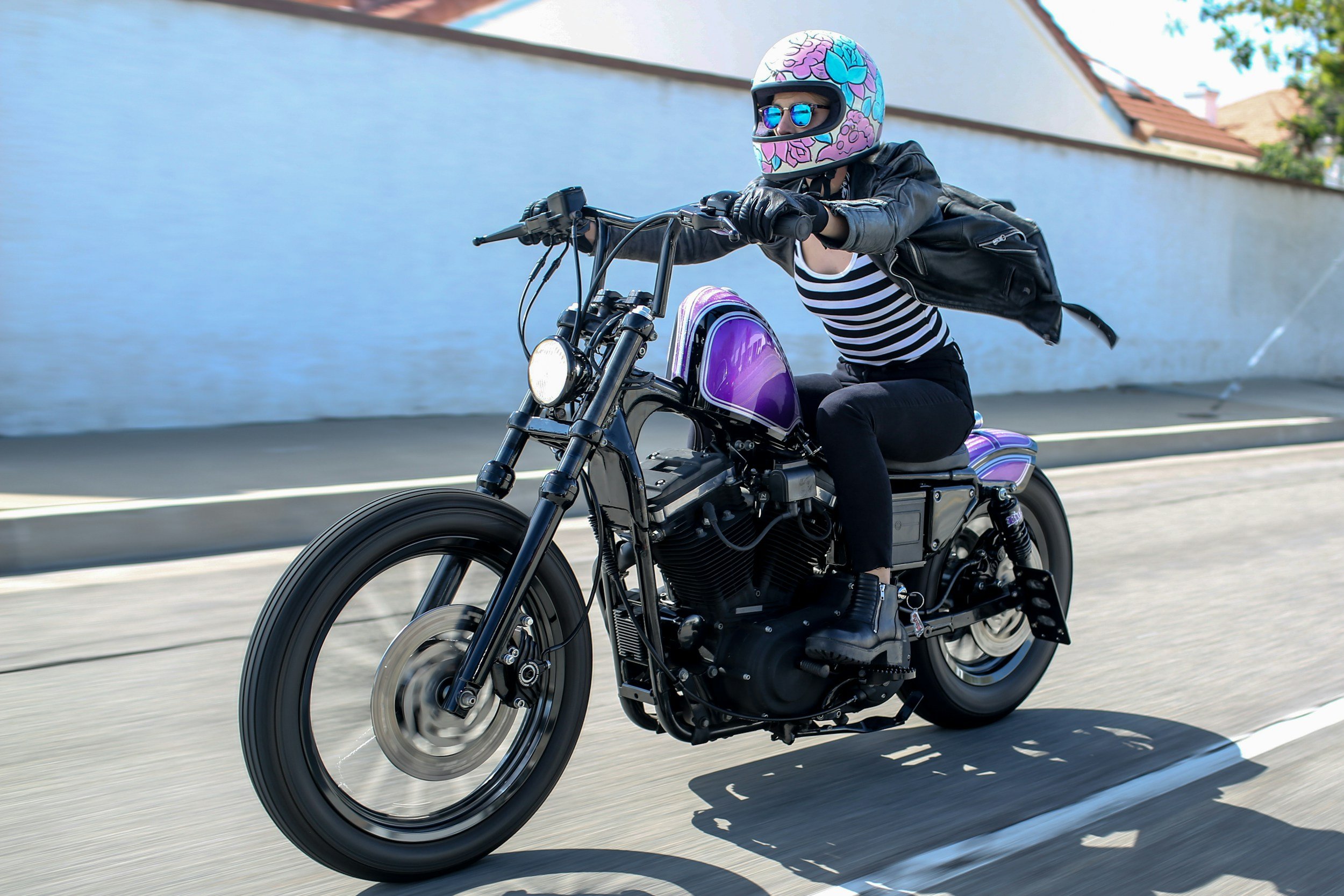 A woman riding a black and purple motorcycle on a street, wearing a floral helmet with sunglasses, a striped shirt, black jeans, and a black leather jacket.