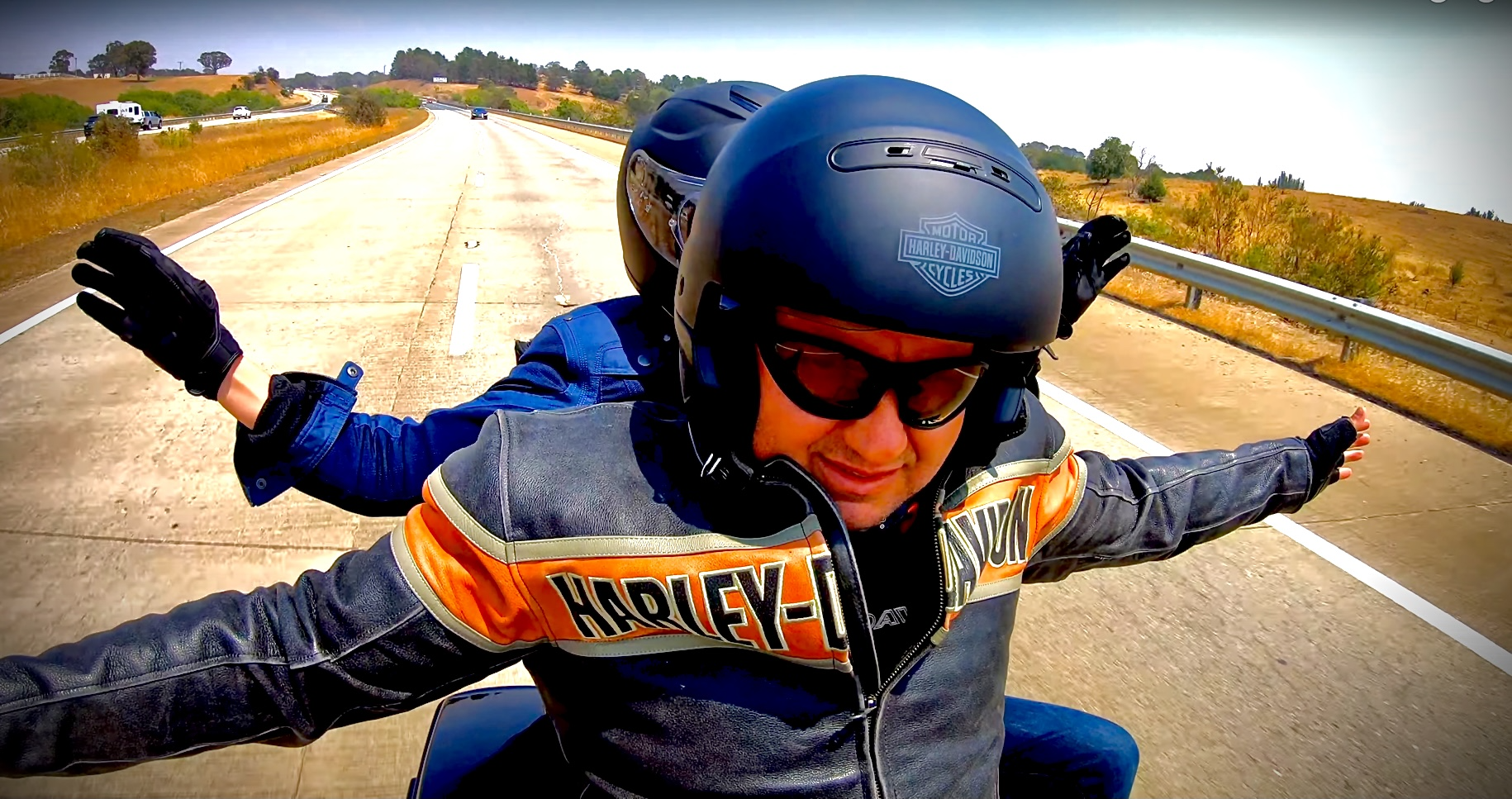 Motorcycle rider and passenger wearing a Harley-Davidson helmet and jacket waving their hands, riding on an open highway with a clear sky and dry landscape.
