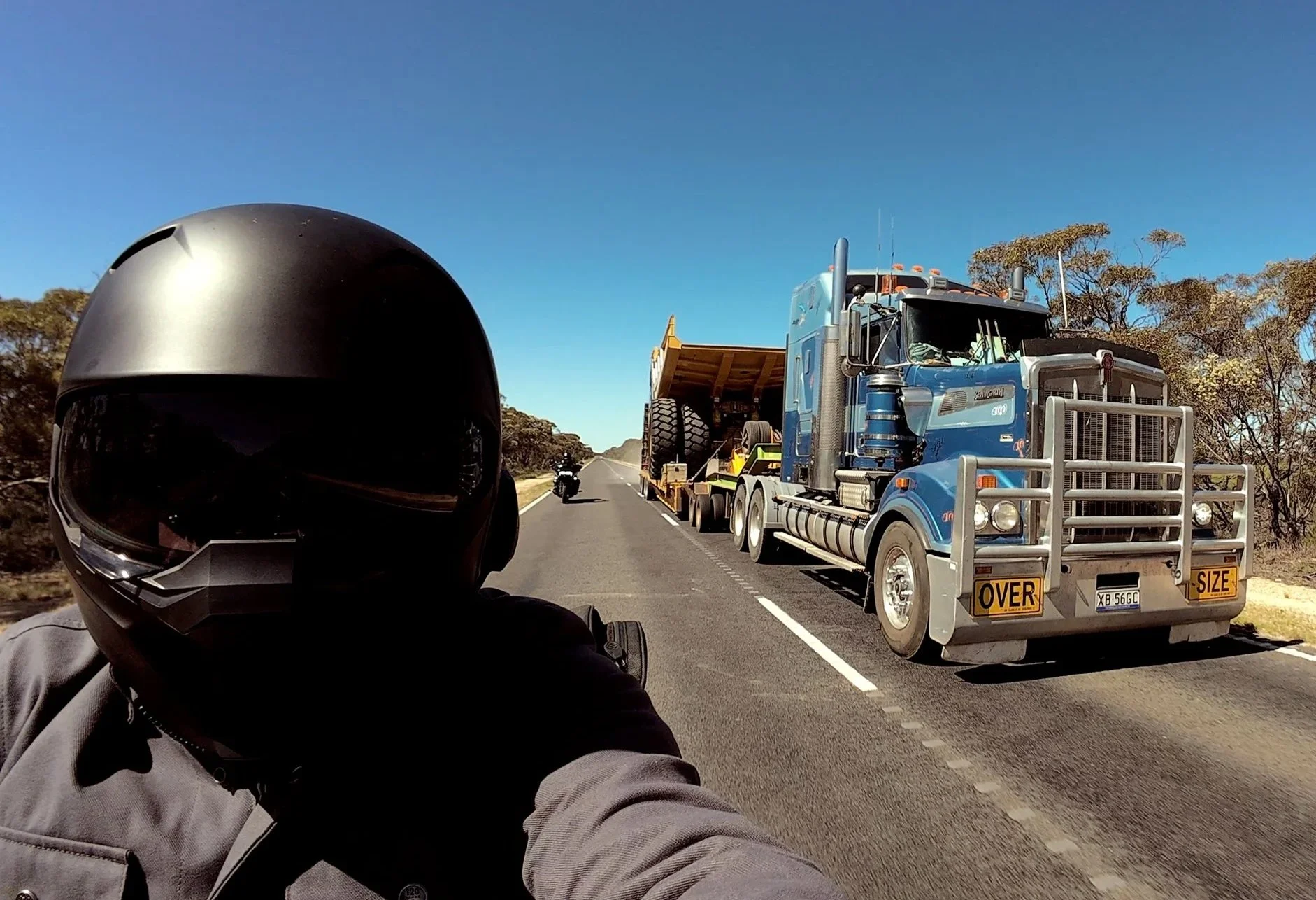 Motorcycle rider overtaking a large semi-truck and other vehicles in the distance, trees lining the side of the road under a blue sky.