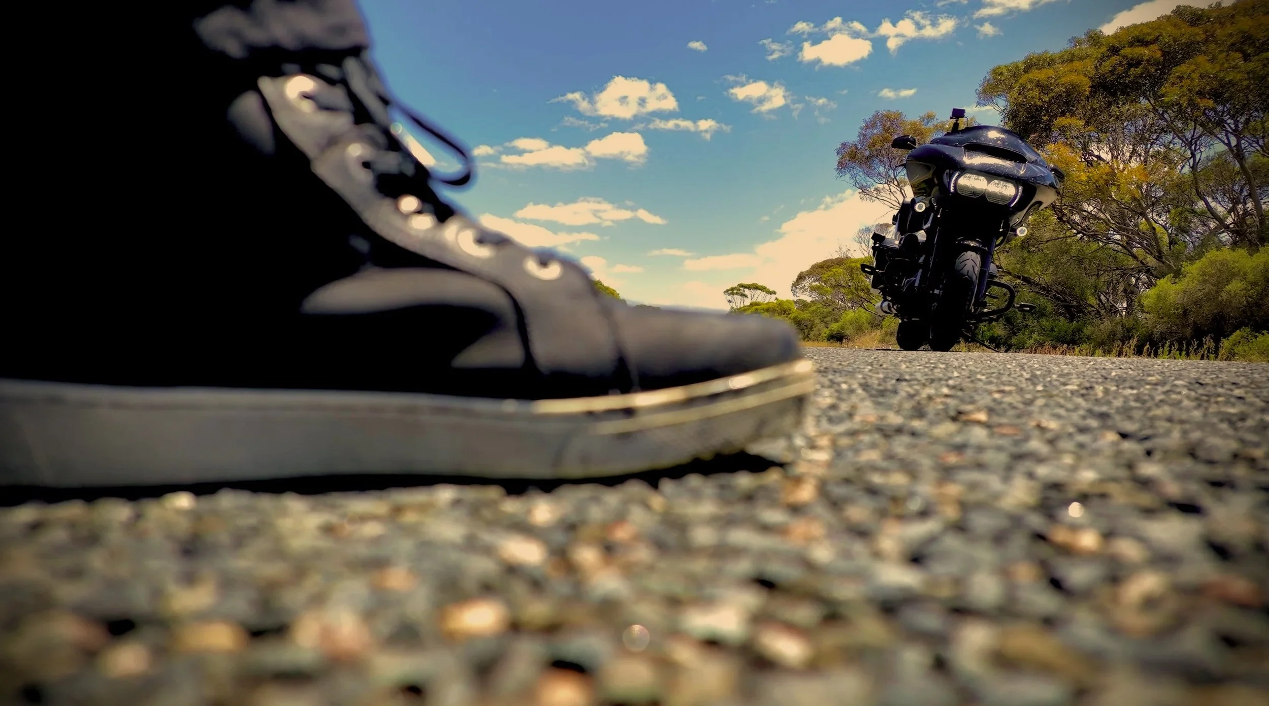 Close-up of a black sneaker on the road with a black motorcycle in the background and a clear blue sky with scattered clouds.