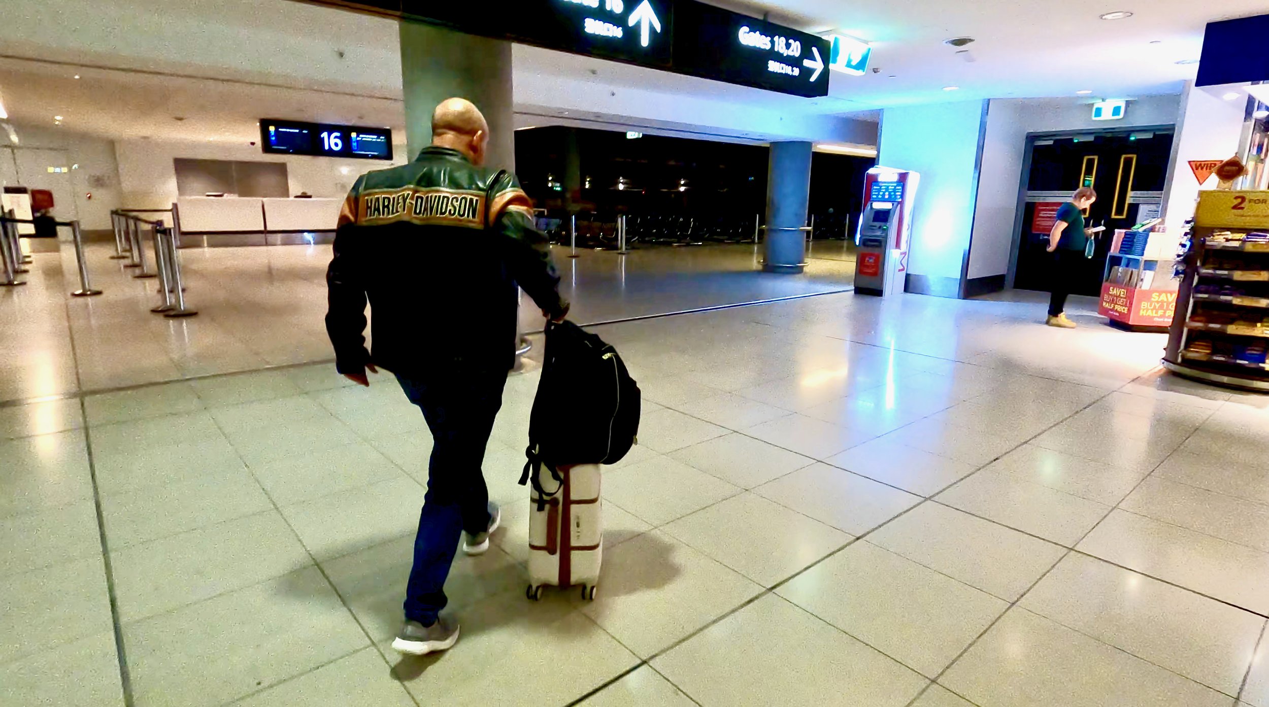 A man in a black Harley Davidson jacket is walking up an escalator at an airport, pulling a rolling suitcase and carrying a backpack, with others in the background.