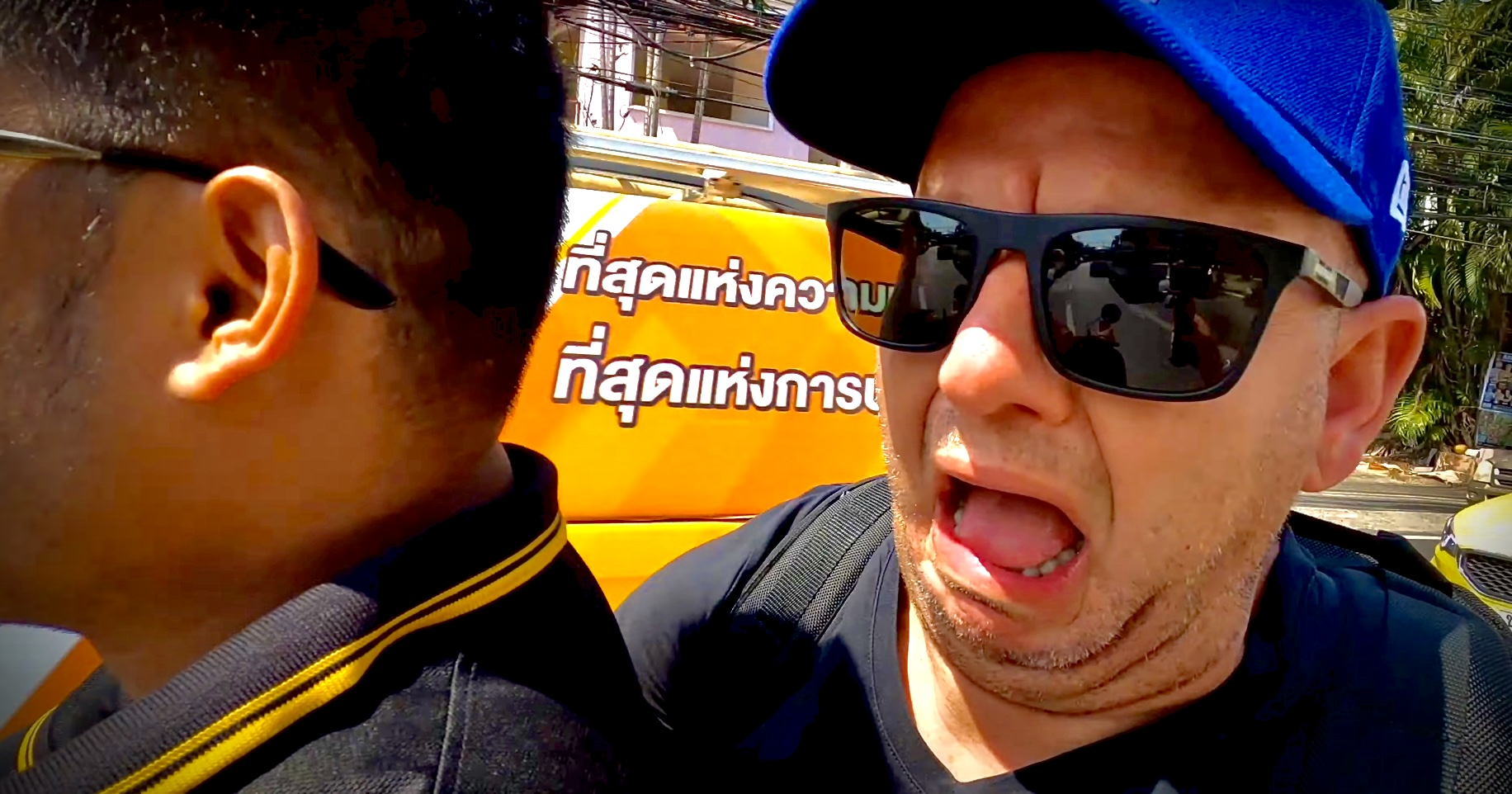 A close-up of a man with sunglasses and a blue cap, appearing to be speaking or yelling at another person, with a yellow sign with Thai text behind him.