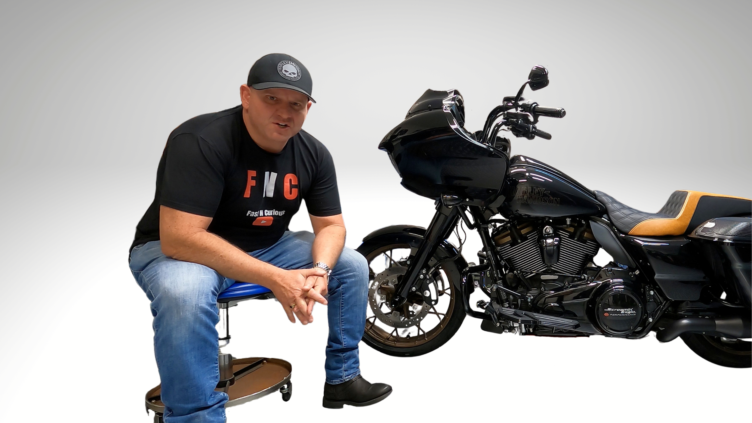 A man wearing a black t-shirt, jeans, and black shoes sitting on a small stool next to a black Harley Davidson motorcycle, with a white background.