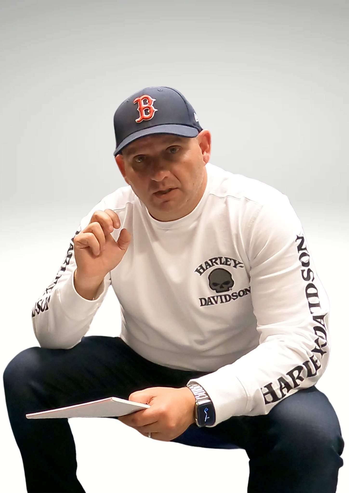 Man wearing a white Harley-Davidson long sleeve shirt and a black baseball cap, crouching with a notepad in his hand and looking at the camera.
