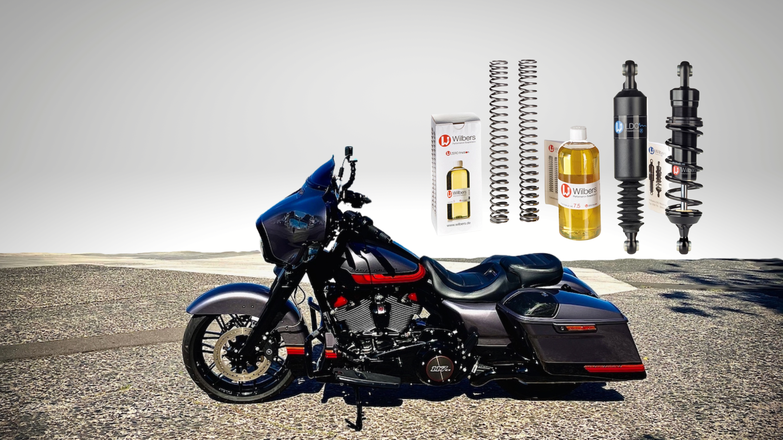 A black and blue touring motorcycle parked on a textured ground with a gradient background. Above the motorcycle, there are various motorcycle suspension parts, including springs, shock absorbers, a bottle of oil, and packaging from Wilbers.