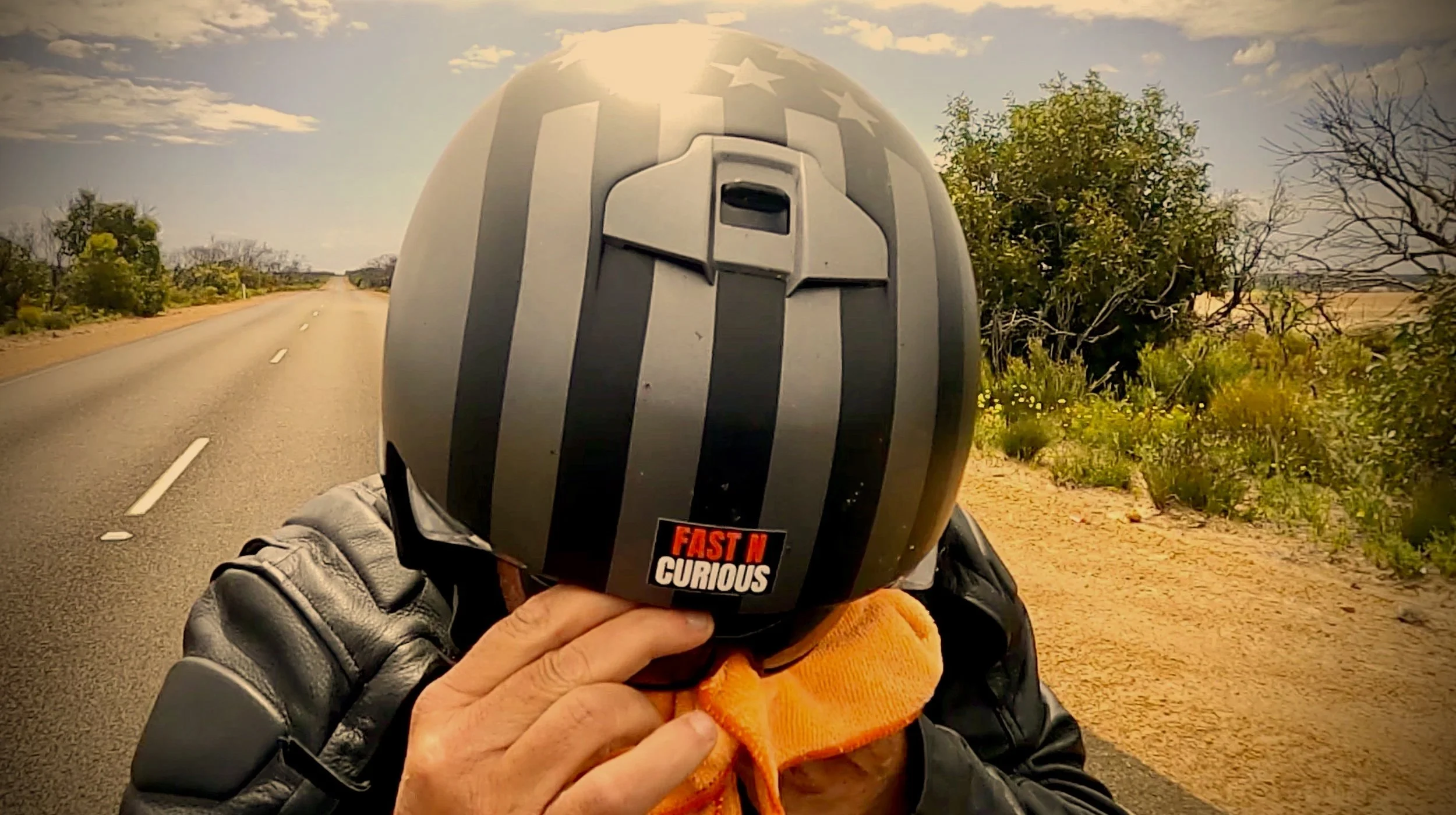 Person wearing a helmet, a black leather jacket, and rubbing their eyes with an orange rag, he is riding down an empty two-lane road with trees and bushes on each side, under a partly cloudy sky.