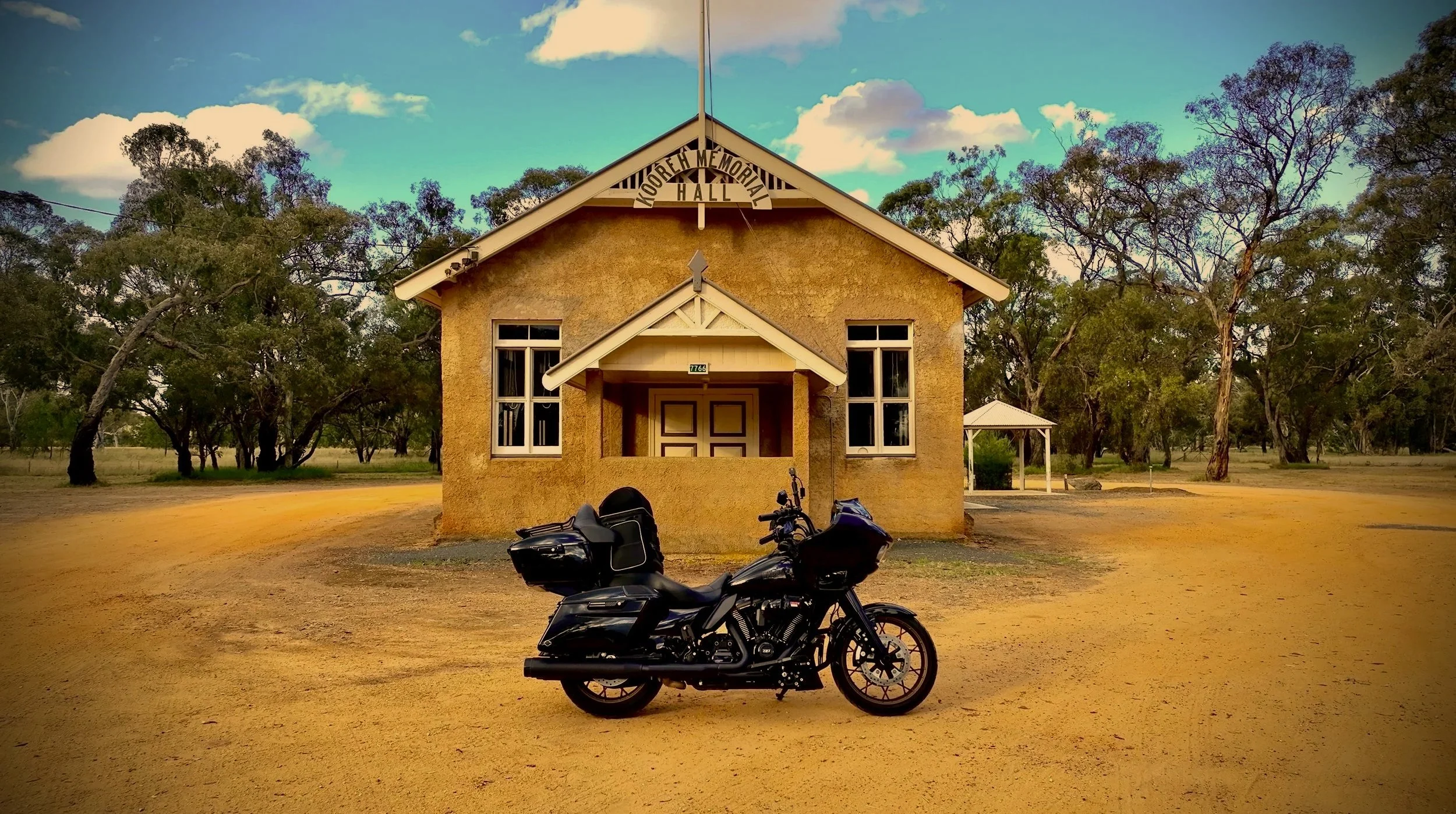 A black motorcycle parked in front of a small, tan-colored wood building with a sign that reads 'Mooreh Memorial Hall' at the top. The building has three windows and a small porch. There are trees and open land surrounding the hall under a partly cloudy sky.