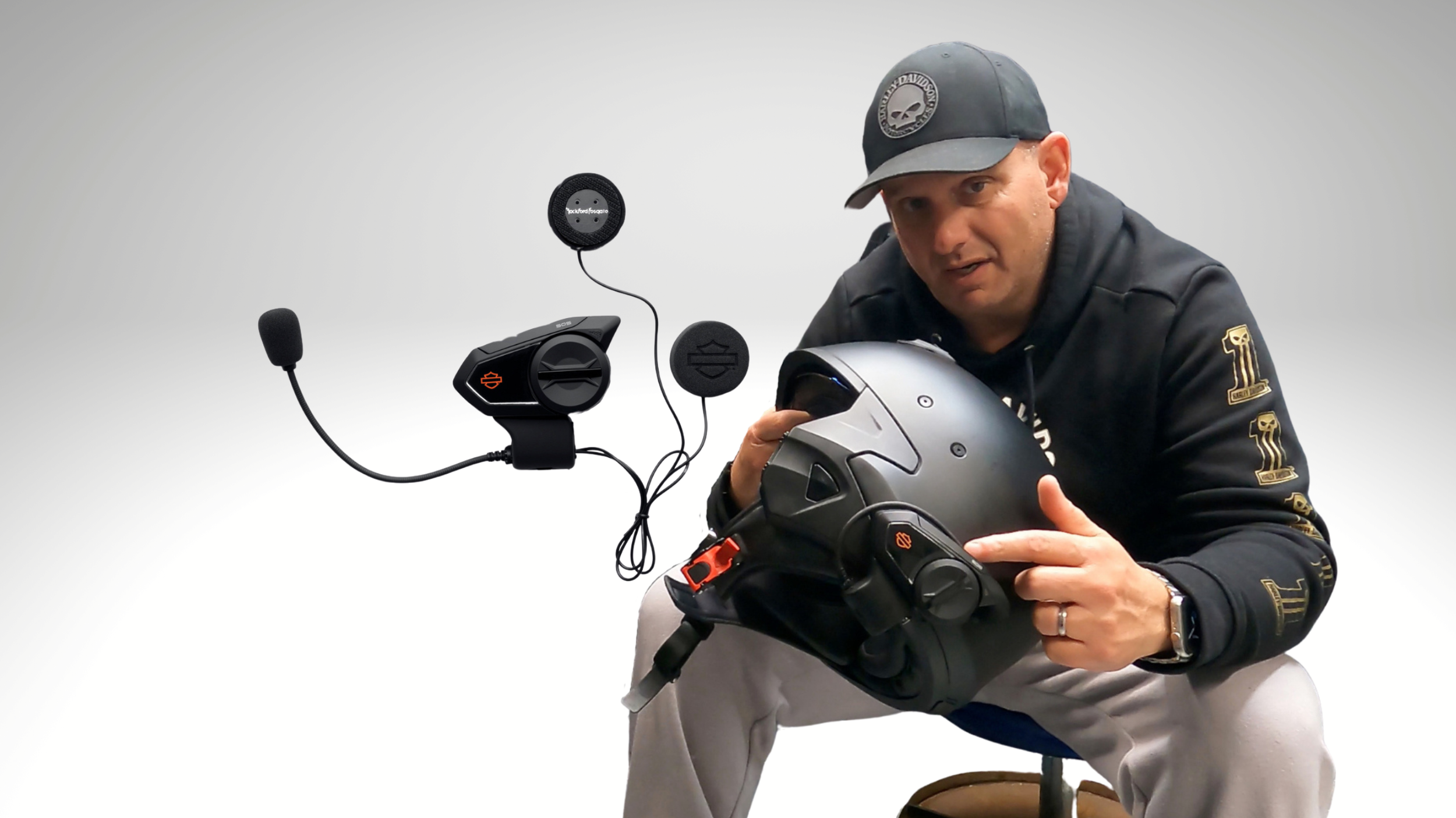 A man sitting on a stool holding a motorcycle helmet with a microphone and communication devices floating nearby.