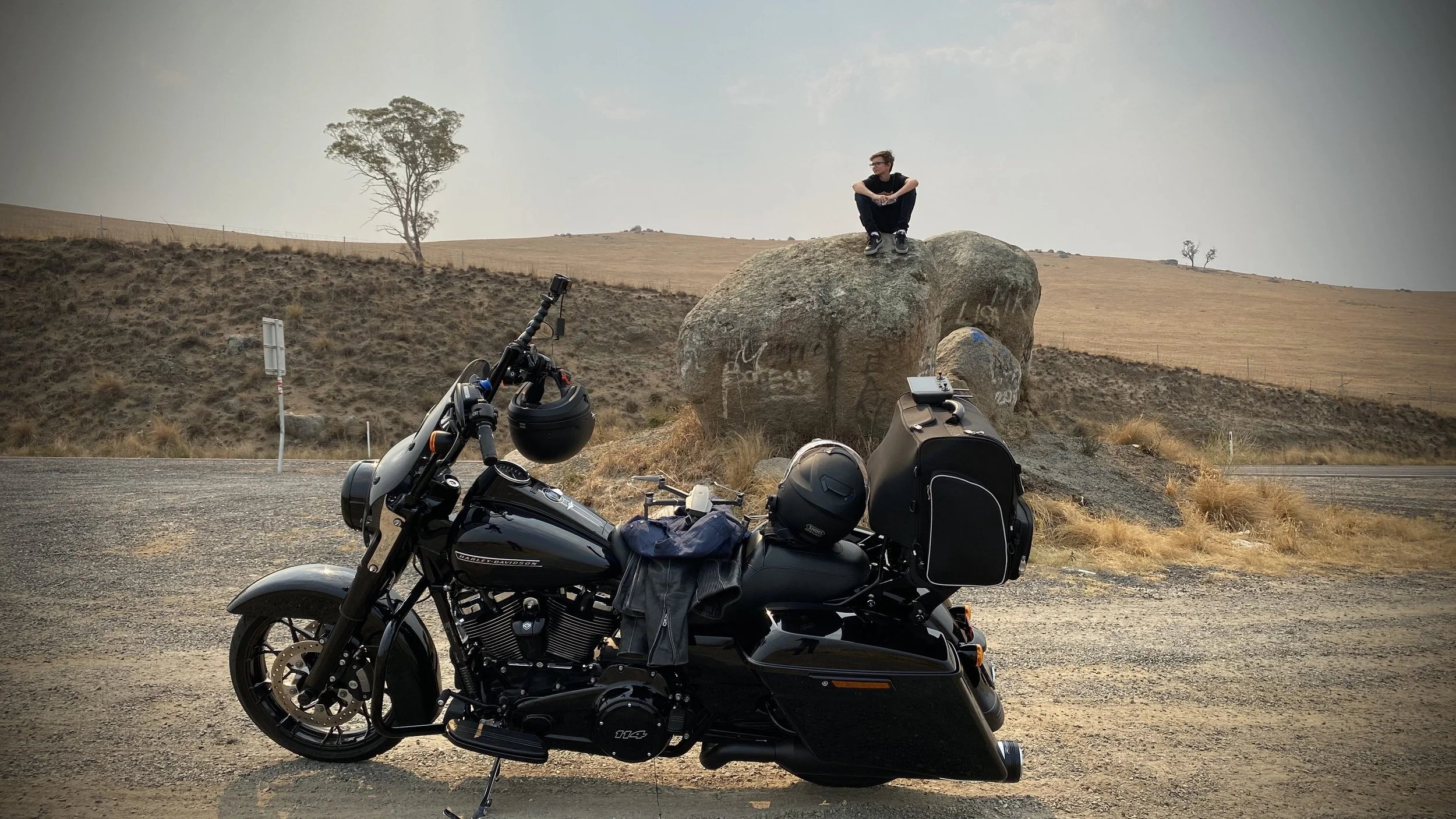 A black motorcycle parked on the side of a rural road with a large boulder in the background. A person is sitting on top of the boulder, looking into the distance, with dry grass and a hillside with scattered trees around.
