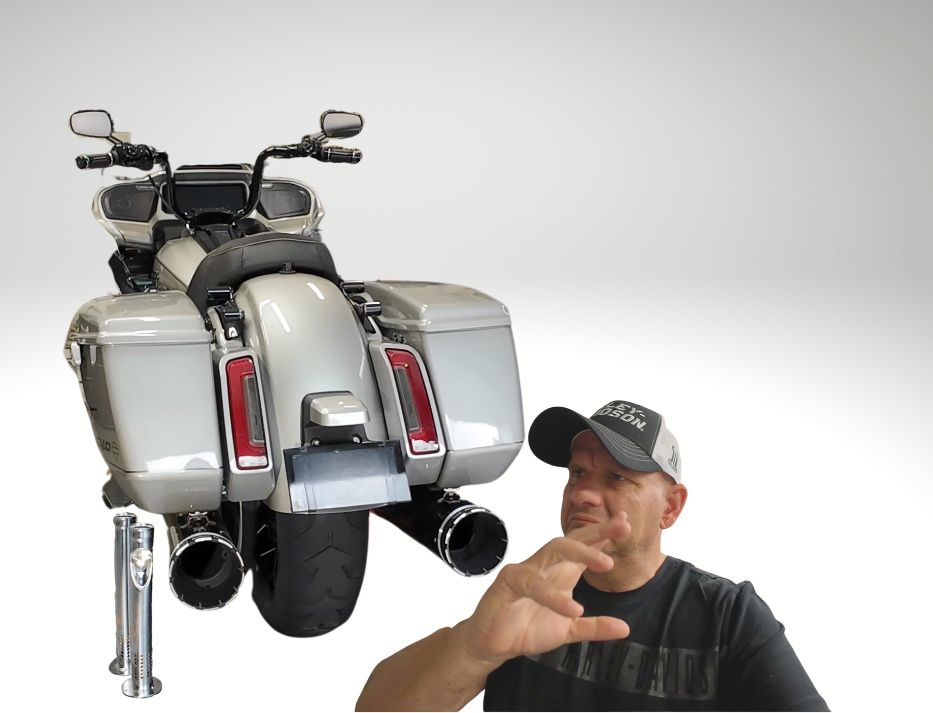 A man wearing a baseball cap and black T-shirt taking a selfie in front of a large, silver motorcycle with saddlebags and a digital display, against a plain white background.