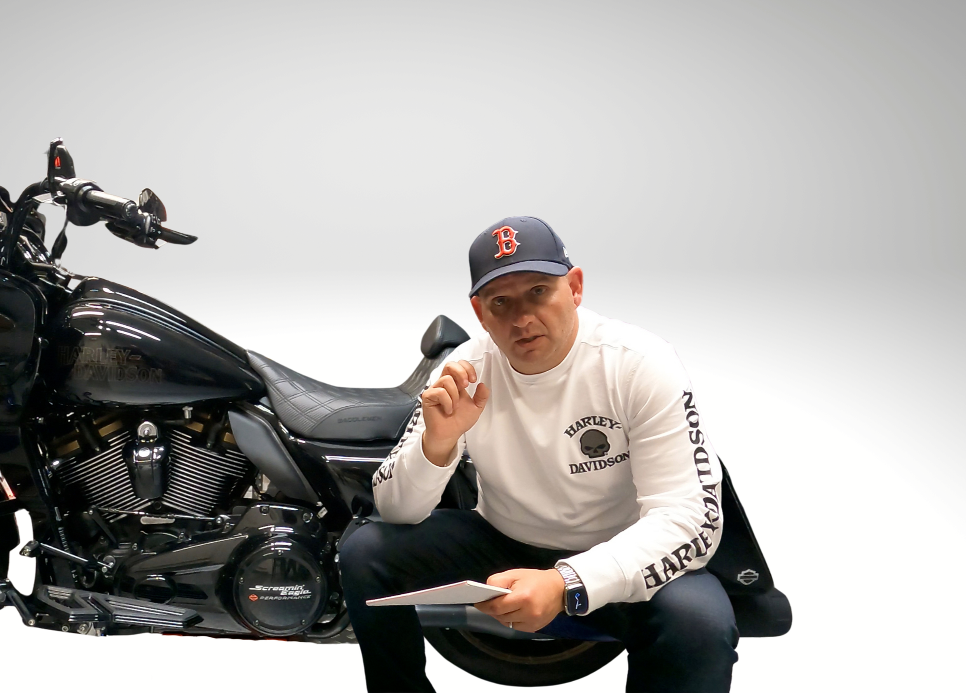 A man wearing a white Harley Davidson sweatshirt and a navy blue cap with a red 'B' logo, kneeling next to a black Harley Davidson motorcycle, holding a notepad and gesturing with his finger against a plain white background.
